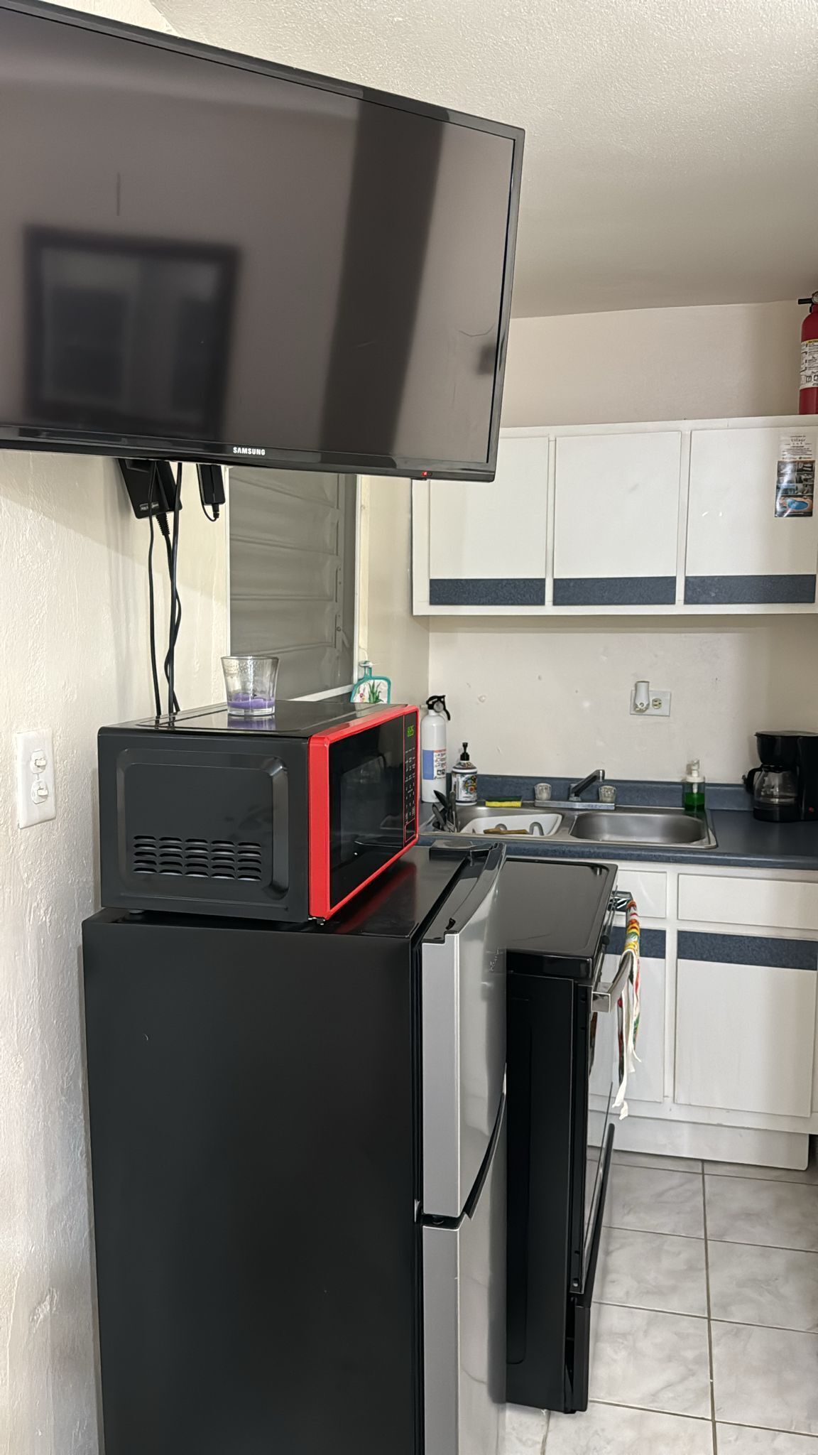 Une cuisine compacte avec un téléviseur à écran plat fixé au-dessus d’un réfrigérateur noir, un four à micro-ondes rouge et un petit évier.