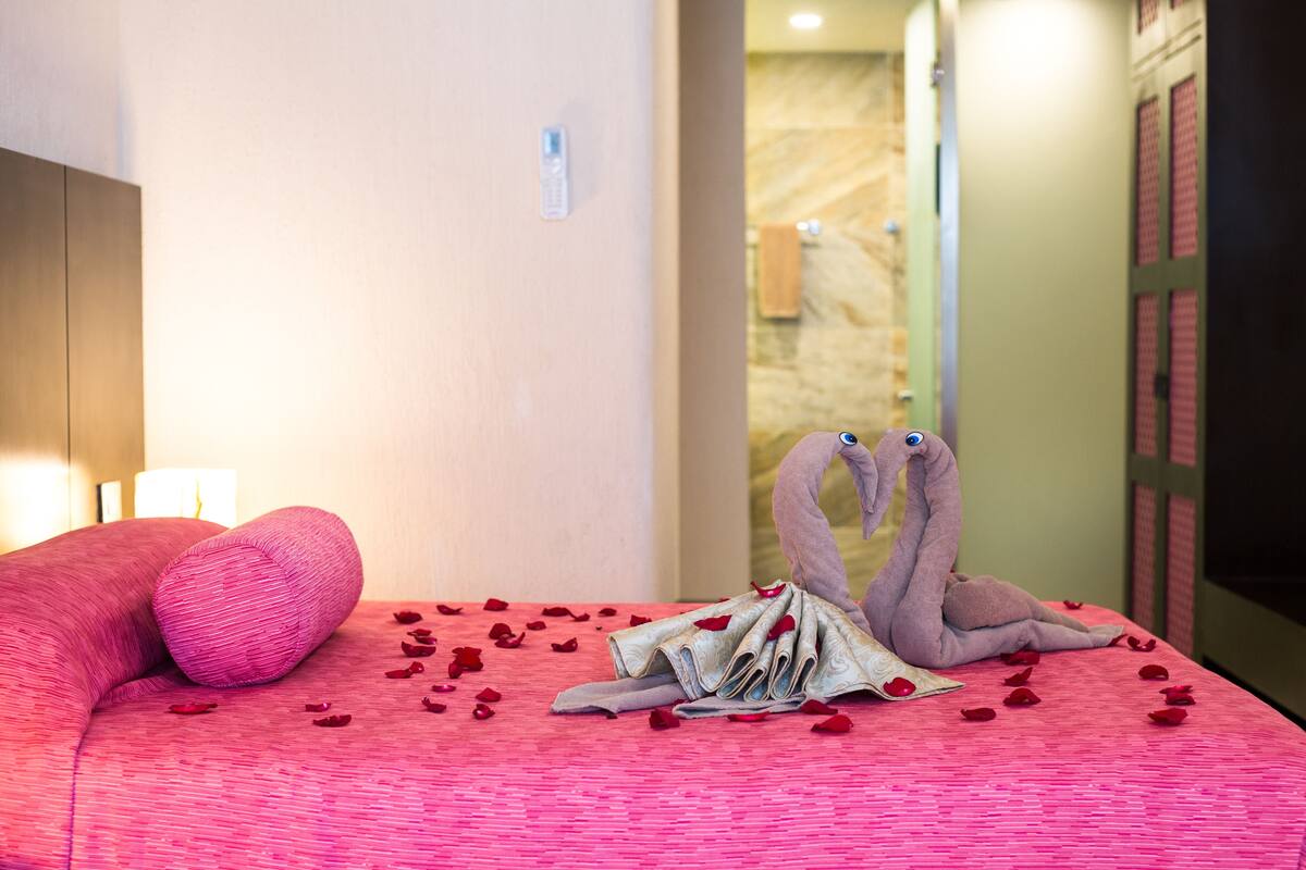 A hotel room with a bed covered in rose petals, a pink pillow, and two swan-shaped towels.