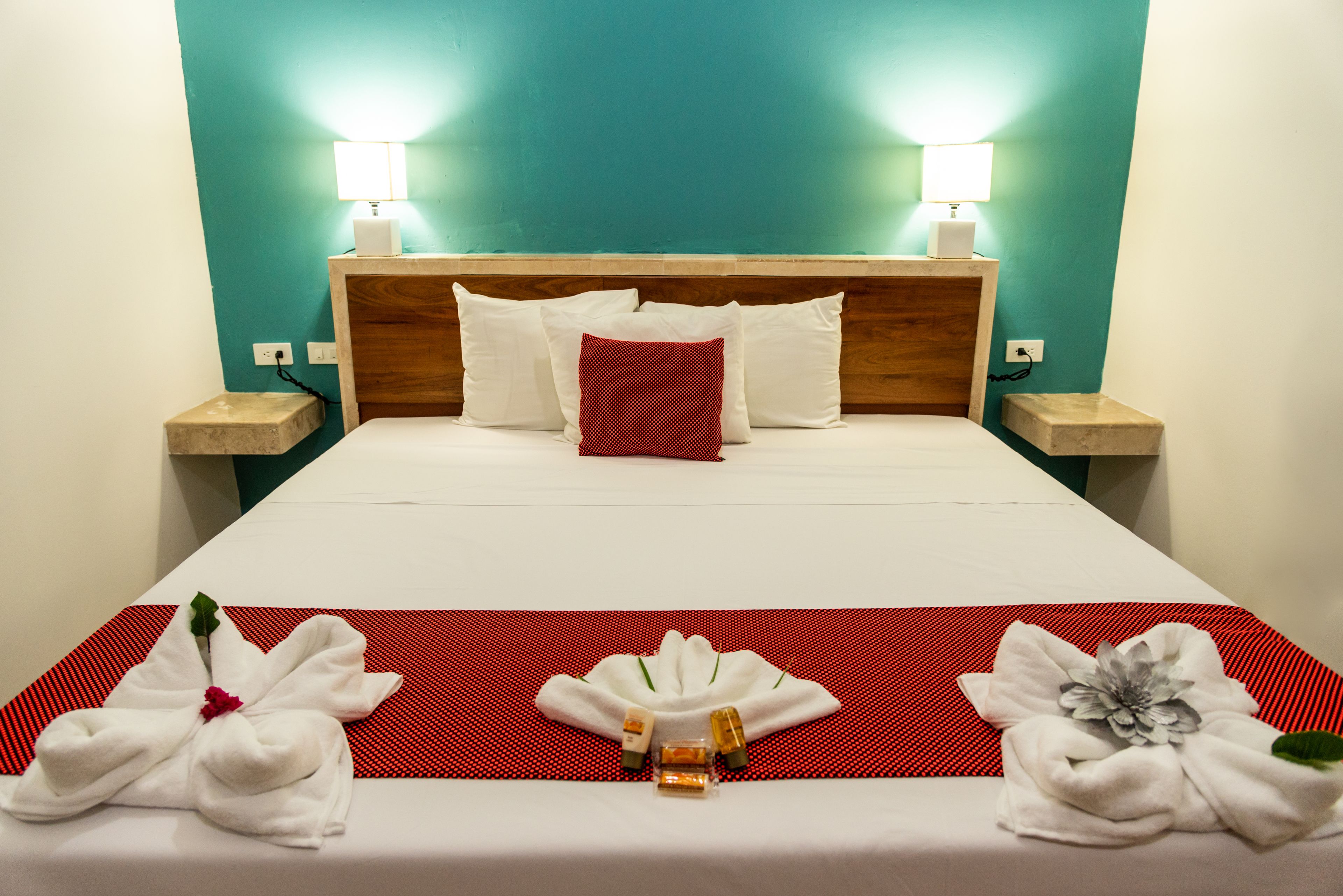 Habitación de hotel con una cama, dos lámparas de noche, una pared color verde azulado y una disposición de toallas rojas y blancas sobre la cama.