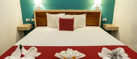 Habitación de hotel con una cama, dos lámparas de noche, una pared color verde azulado y una disposición de toallas rojas y blancas sobre la cama.