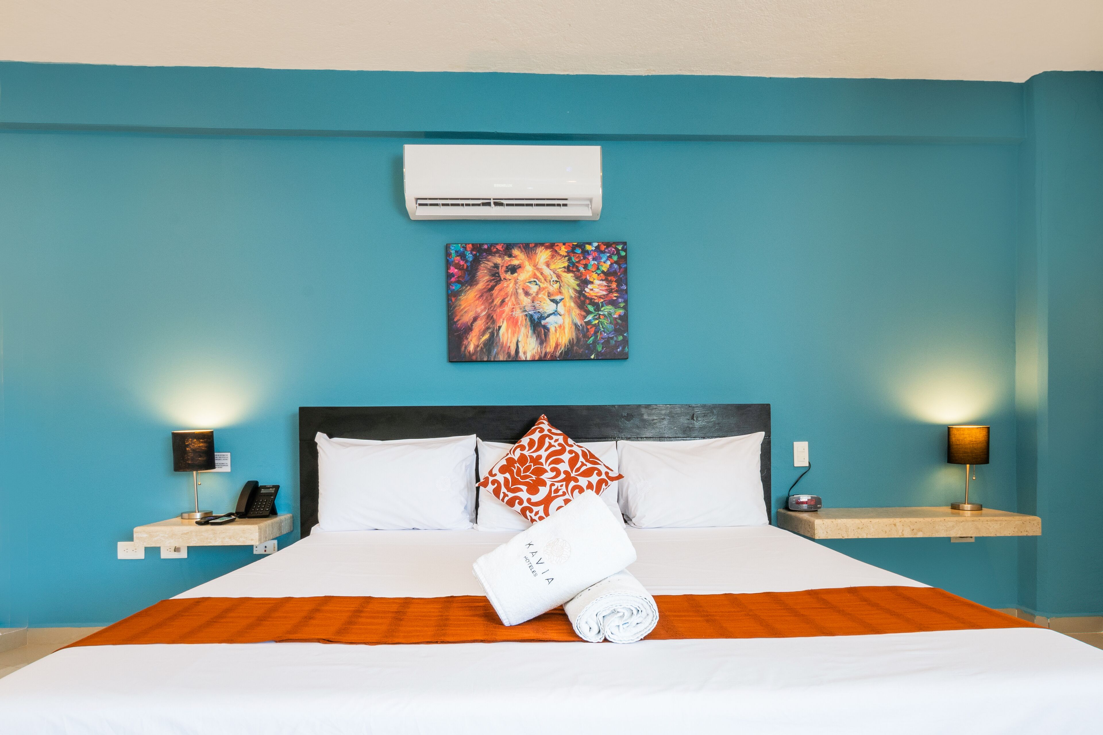 A hotel room with a bed, a painting above the bed, a nightstand with a lamp, and a wall-mounted air conditioner.