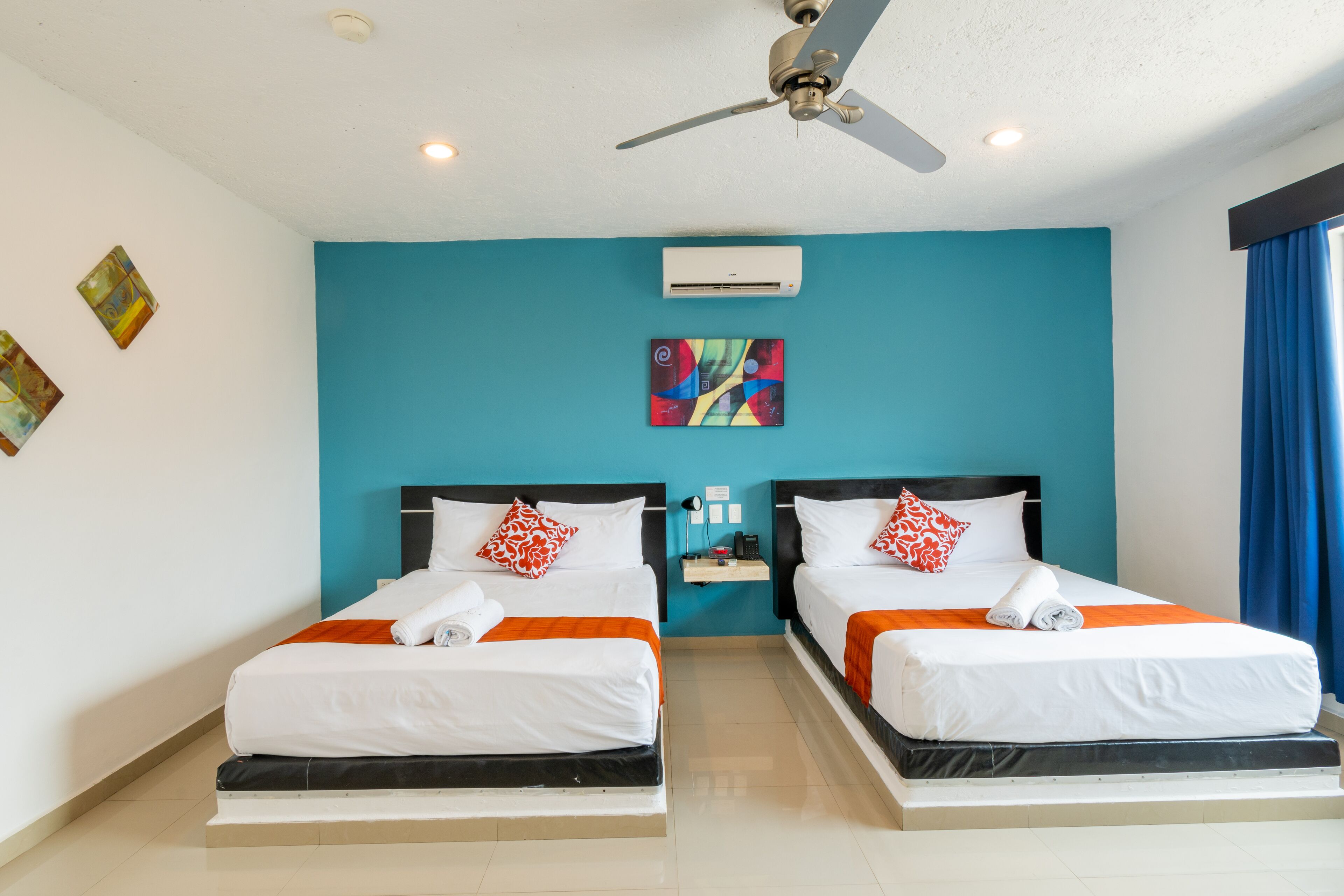 A hotel room with two beds, a ceiling fan, an air conditioning unit, and artwork on the walls.