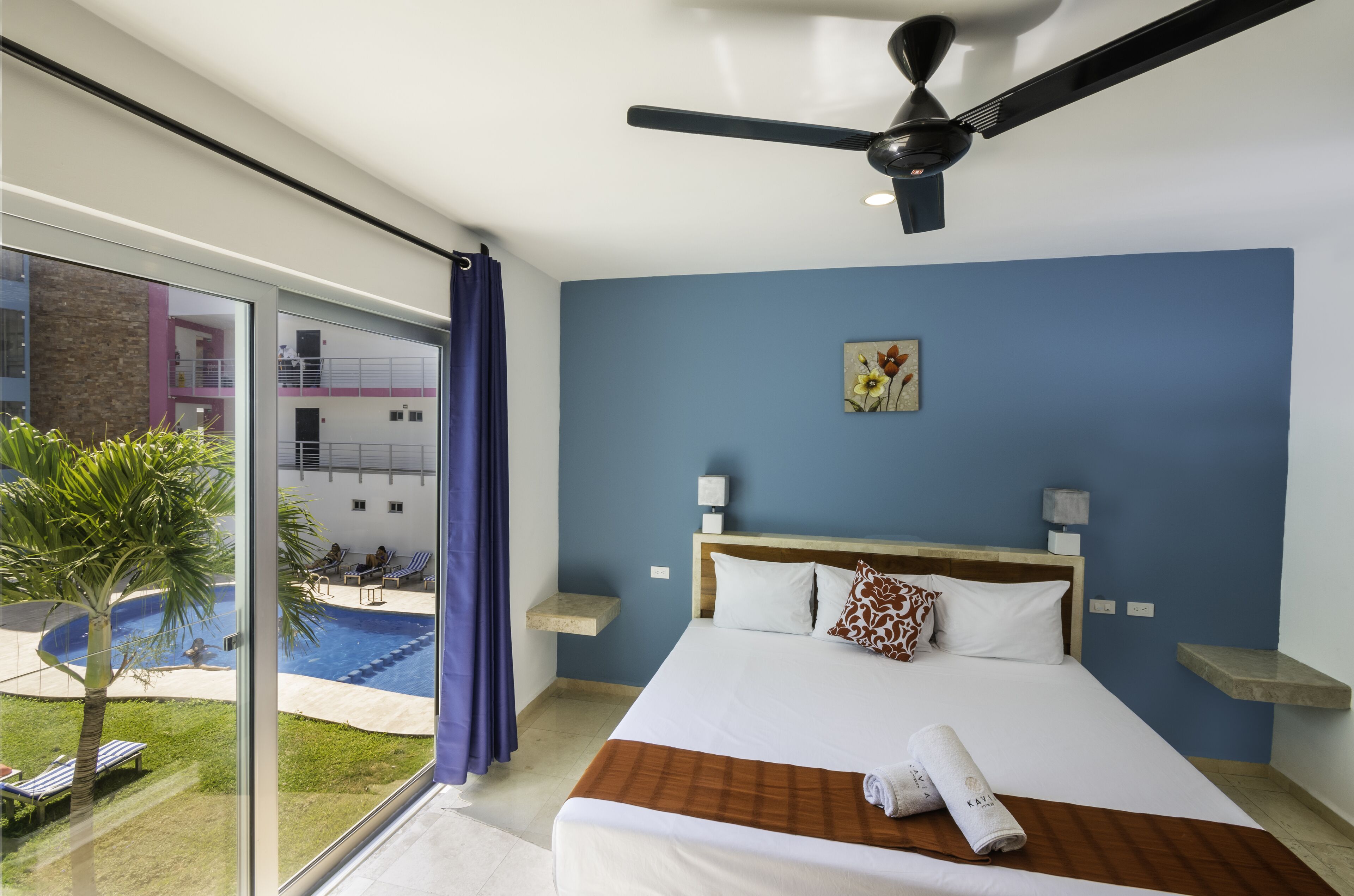 A hotel room with a large bed, a ceiling fan, a view of a swimming pool, and a balcony with a palm tree.