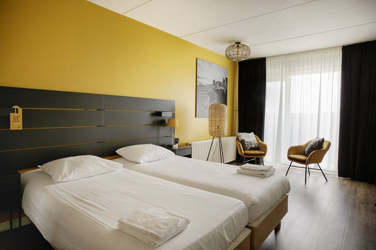 Habitación de hotel con dos camas, cabecero, una silla, una lámpara y una ventana con cortinas.
