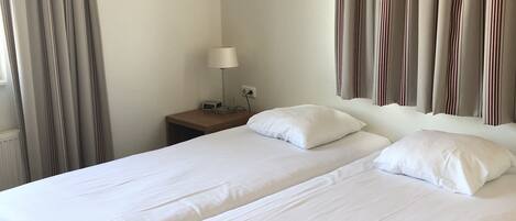 A hotel room with two beds, a nightstand with a lamp, and curtains.