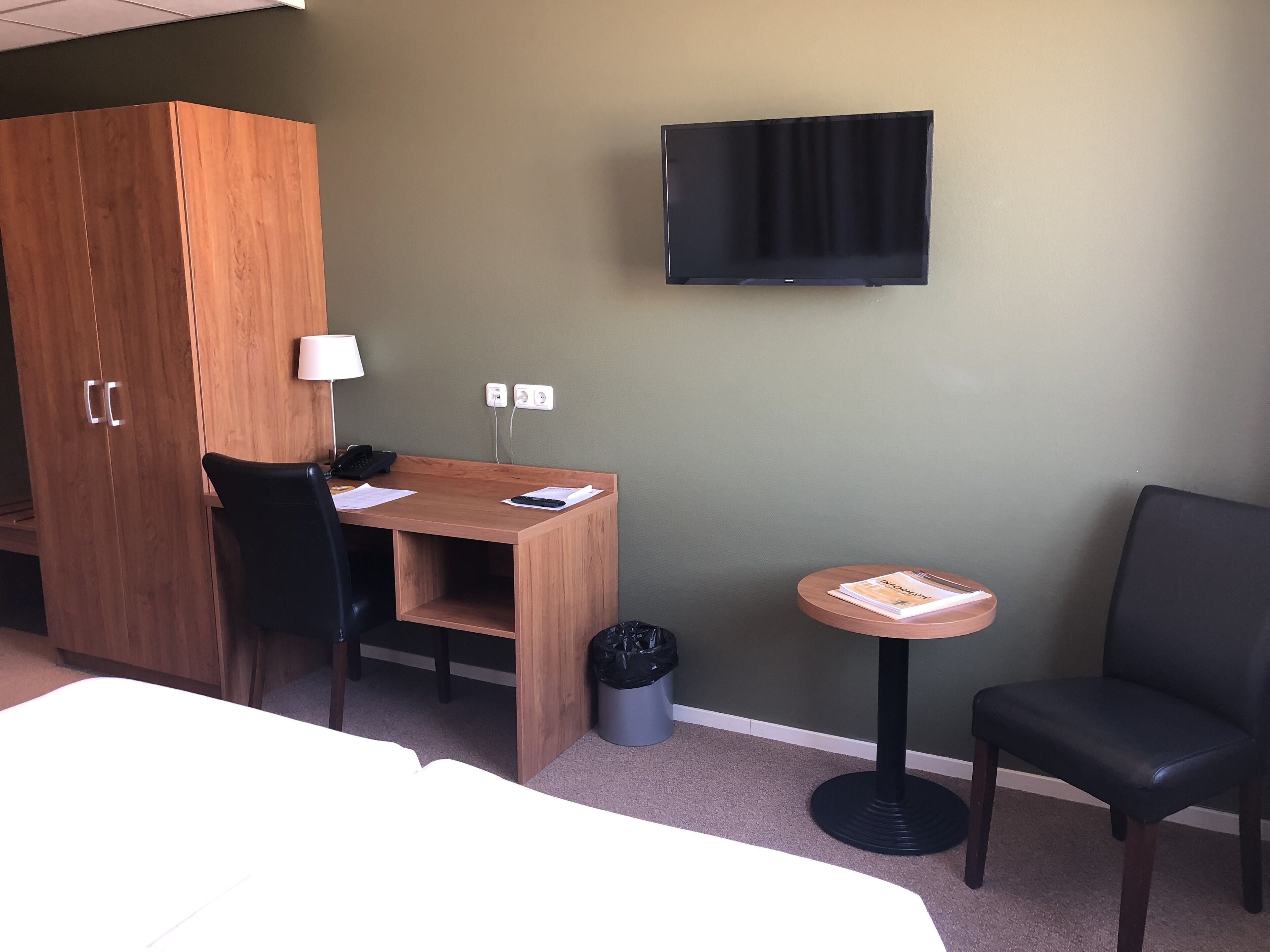 Habitación de hotel con un escritorio de madera, una silla, una mesita redonda con un libro, un sillón de cuero negro, un televisor montado en la pared y un armario.