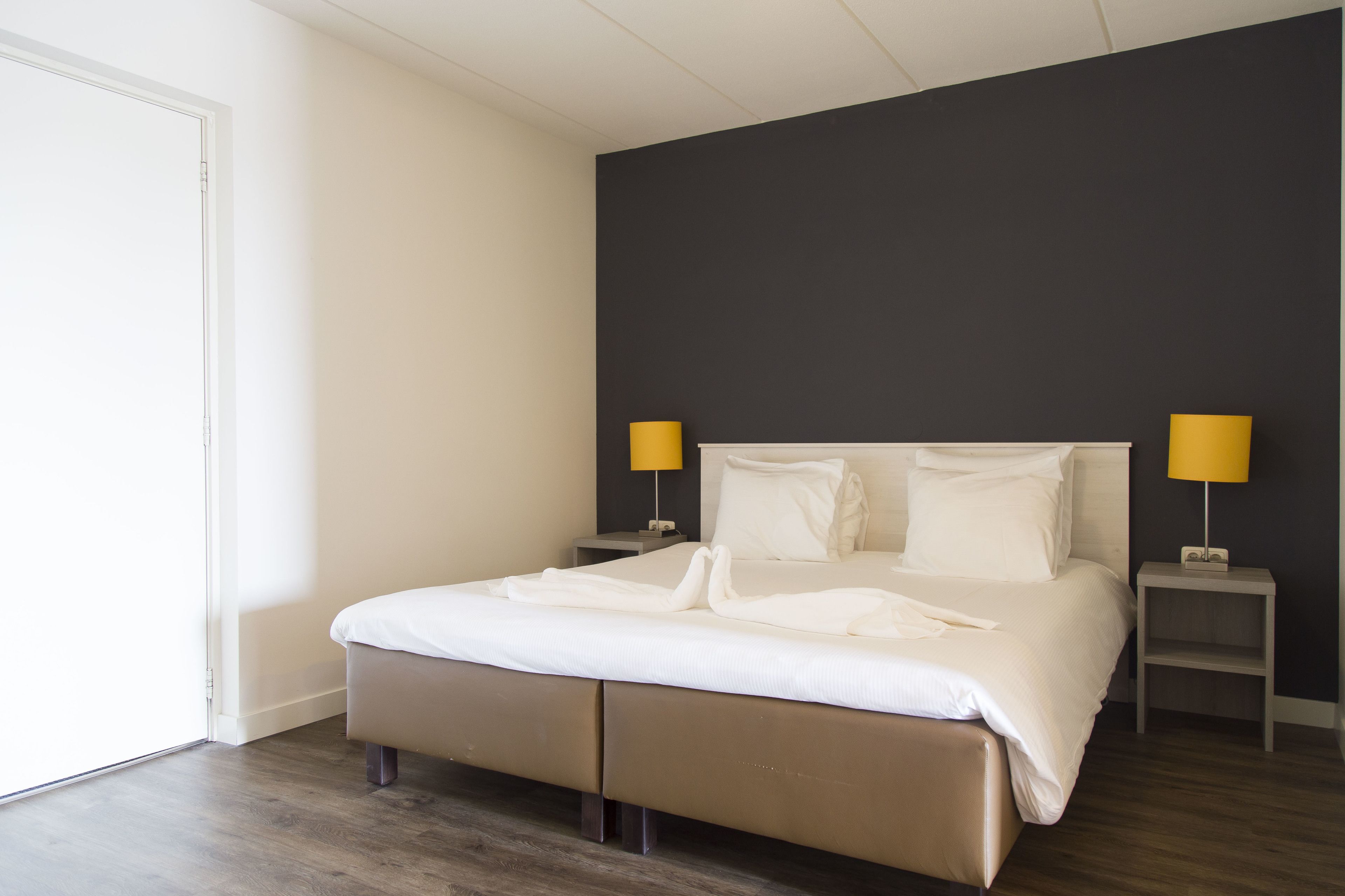 Een net opgemaakt bed met witte lakens, twee nachtkastjes met lampen, en een donkere accentmuur.