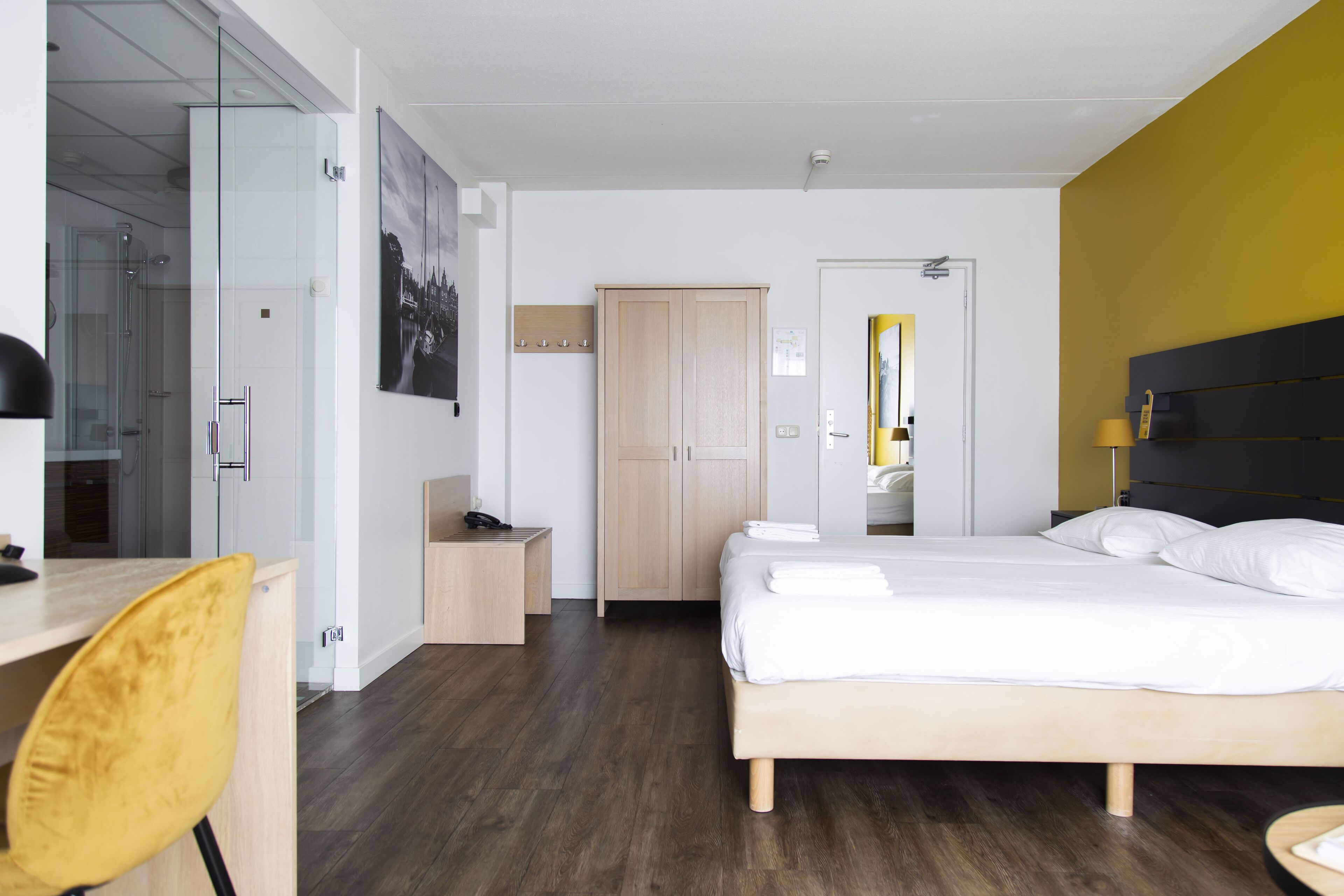 A modern hotel room with a wooden bed, a desk with a chair, a wardrobe, and a bathroom visible through a glass door.