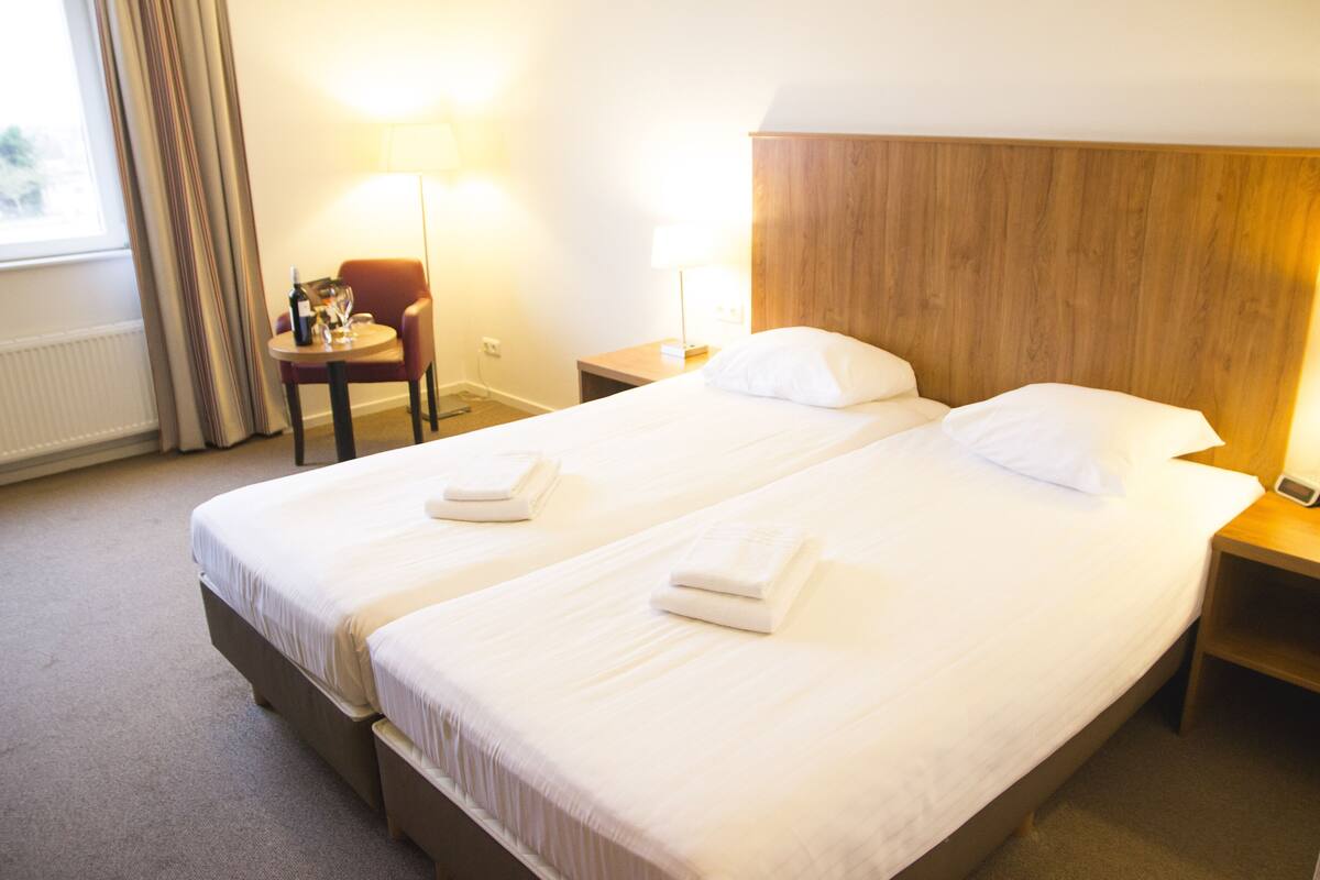 Habitación de hotel con dos camas, una mesita pequeña con una silla, una lámpara y una mesita de noche con un teléfono.
