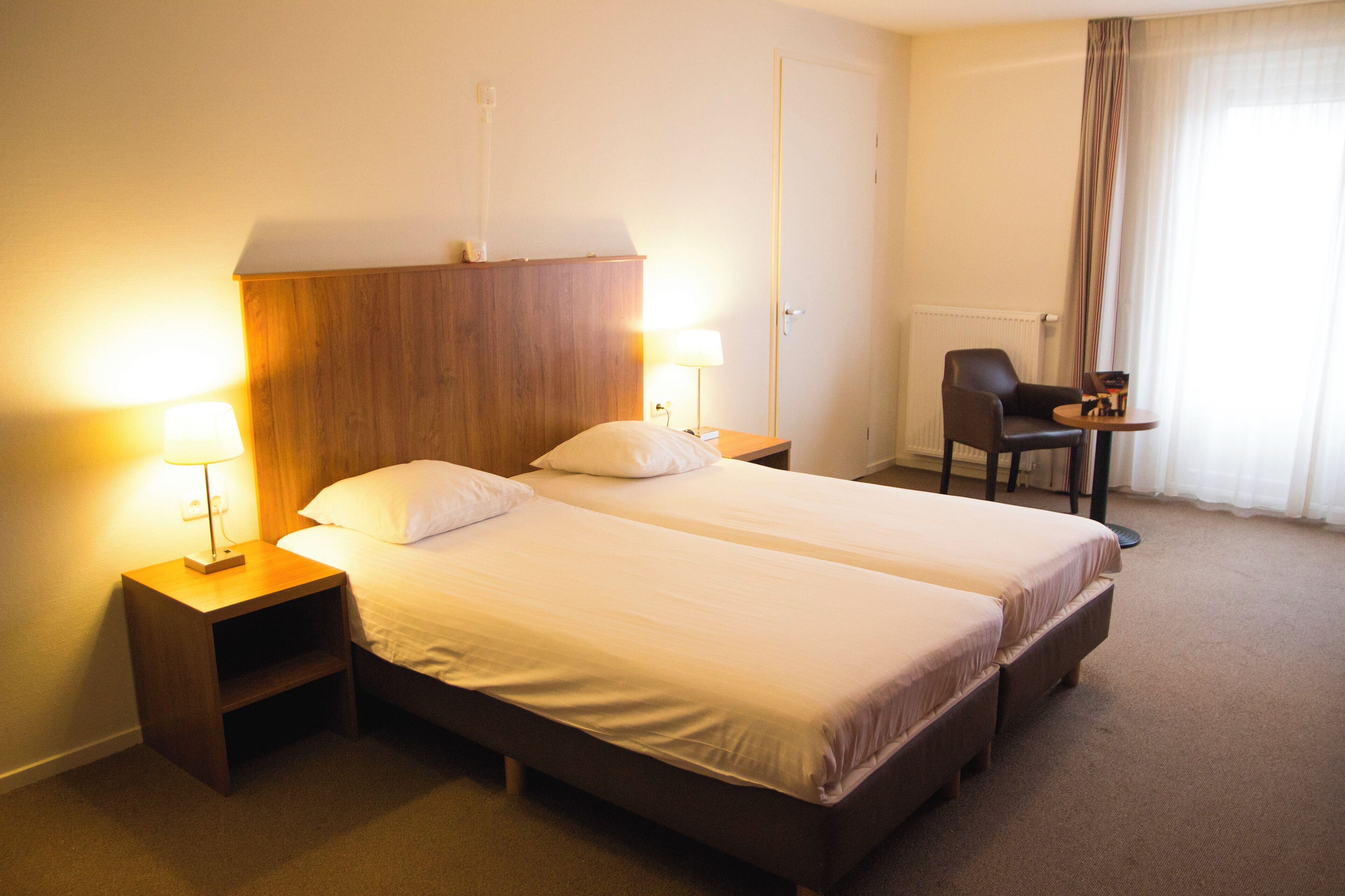 A hotel room with two single beds, bedside lamps, a wooden headboard, a desk with a chair, and a window with curtains.