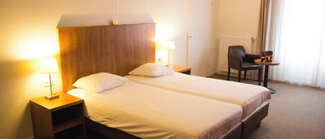 Quarto de hotel com duas camas de solteiro, abajures, cabeceira de madeira, escrivaninha com cadeira e janela com cortinas.