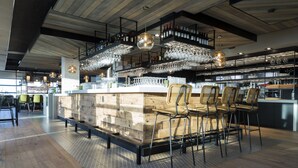 Una zona de bar con mostrador de madera, varios taburetes, lámparas colgantes y estanterías llenas de botellas y vasos.
