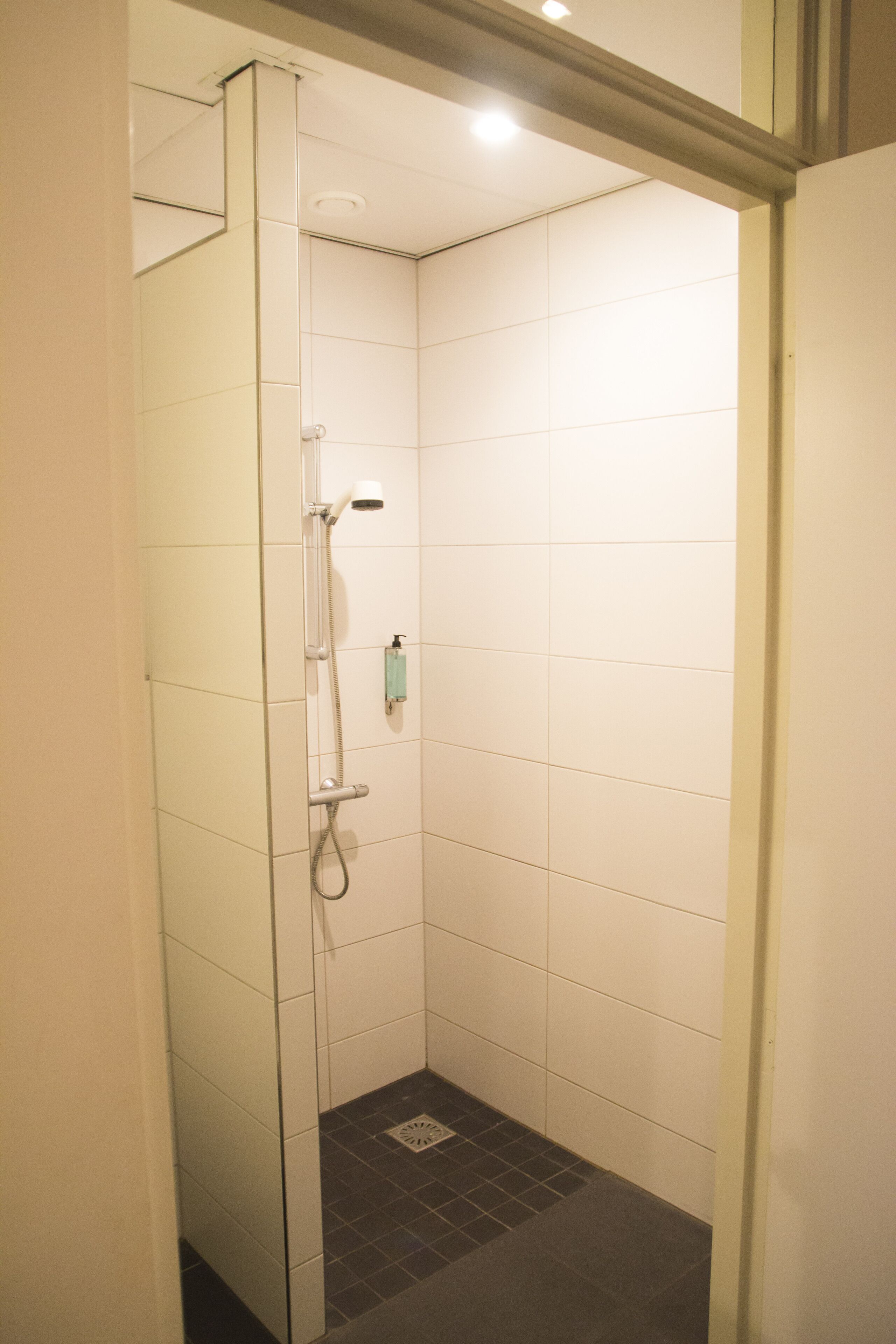 A small, enclosed shower area with white tiles and a glass door, equipped with a handheld showerhead and a handheld shower attachment.