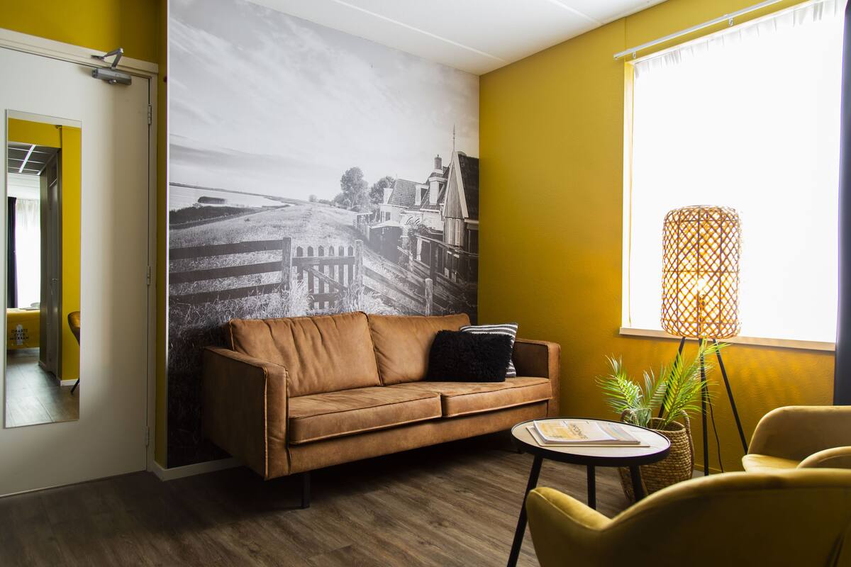 Una habitación con un sofá de cuero marrón, una mesita redonda pequeña, una planta en maceta y un gran mural blanco y negro en la pared.