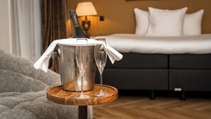 Habitación de hotel con cama, mesita de noche con cubo para champán y copas, lámpara y un cuadro en la pared.