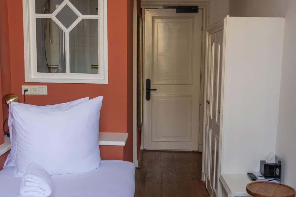 Habitación de hotel con una cama, suelo de madera, un armario blanco, una pequeña mesa de madera y una puerta que da a otra habitación.