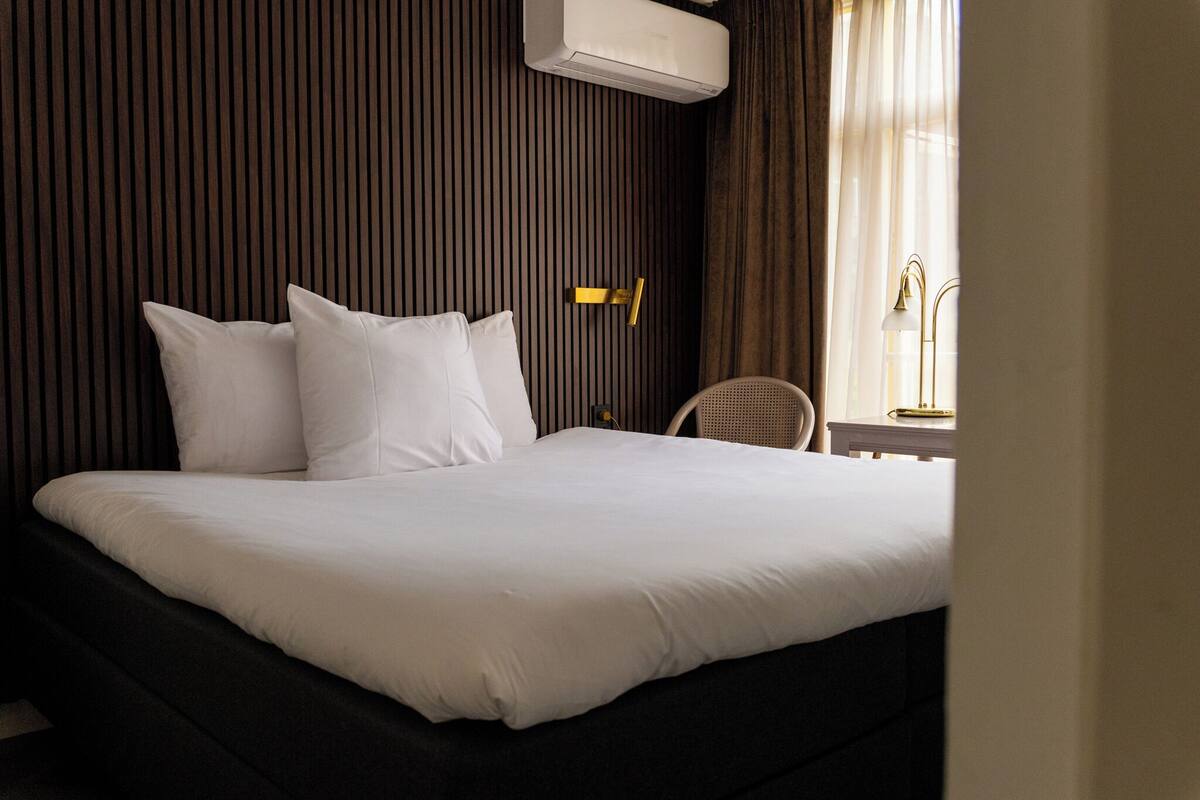 Habitación de hotel con una cama grande, una mesita de noche, una silla y un ventanal con cortinas.