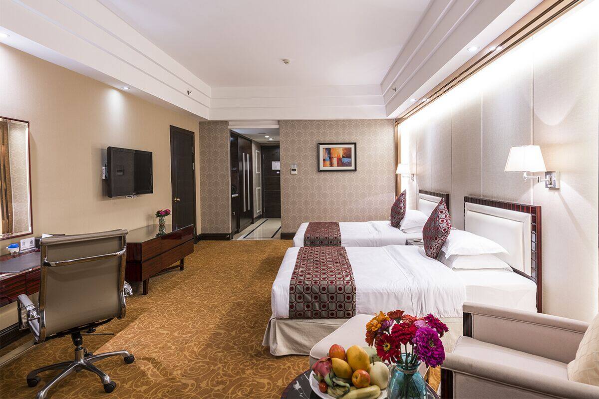 Ein Hotelzimmer mit zwei Betten, einem Schreibtisch mit Stuhl, einem Fernseher und einem Esstisch mit Obst und Blumen.