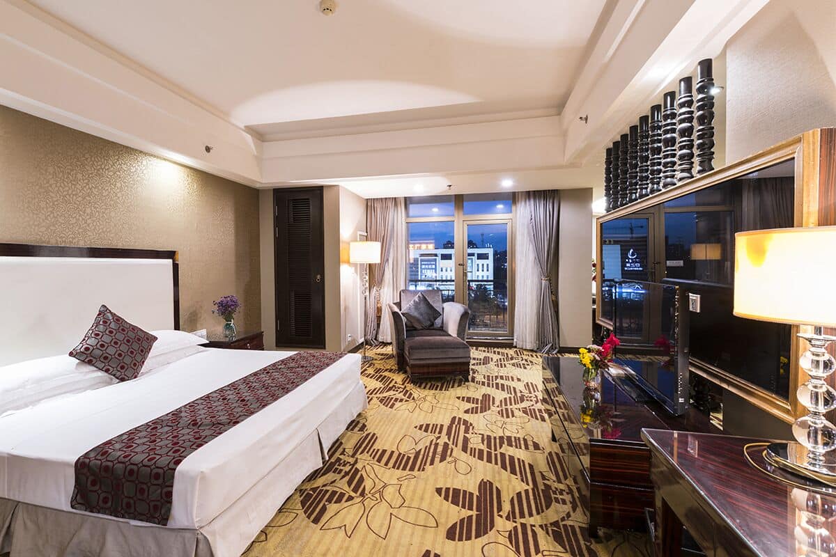 Ein Hotelzimmer mit einem großen Bett, einem Schreibtisch, einem Stuhl, einem Fernseher und Blick auf die nächtliche Stadt.