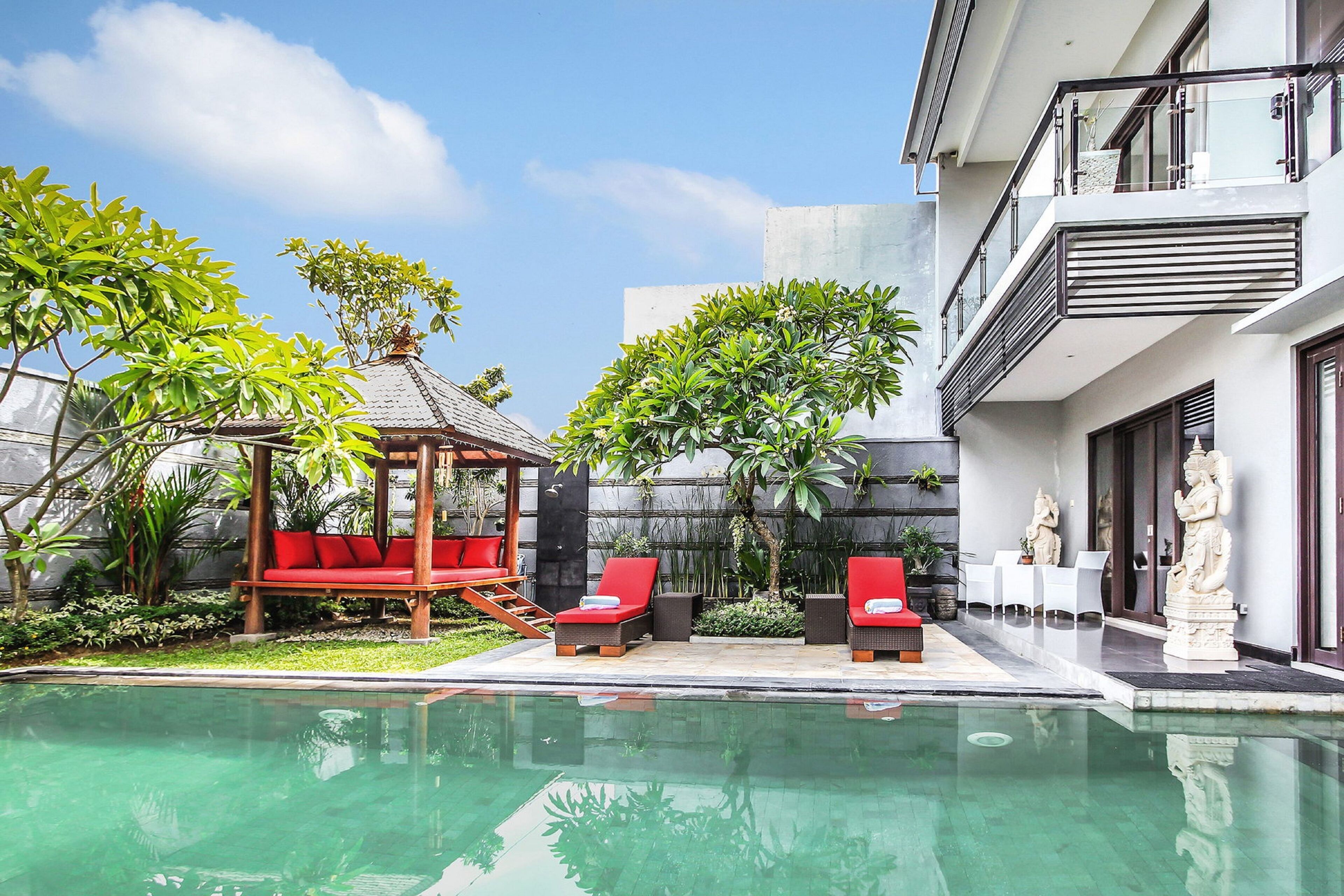 A modern outdoor pool area with red lounge chairs, a wooden gazebo, and a statue.