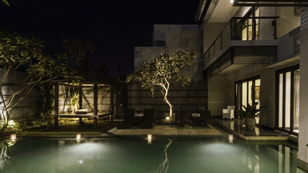 Un área de piscina moderna al aire libre con un árbol, sillas de descanso y un patio bien iluminado.