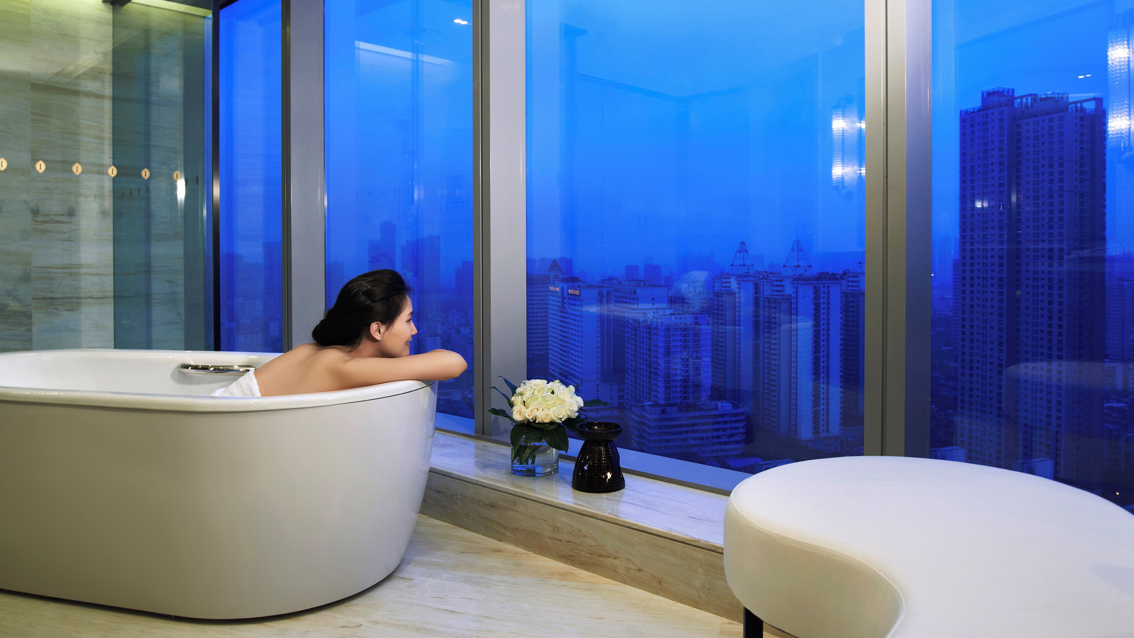 Une salle de bain moderne avec une baignoire, une fenêtre donnant sur la ville et une petite table sur laquelle est posé un vase de fleurs.