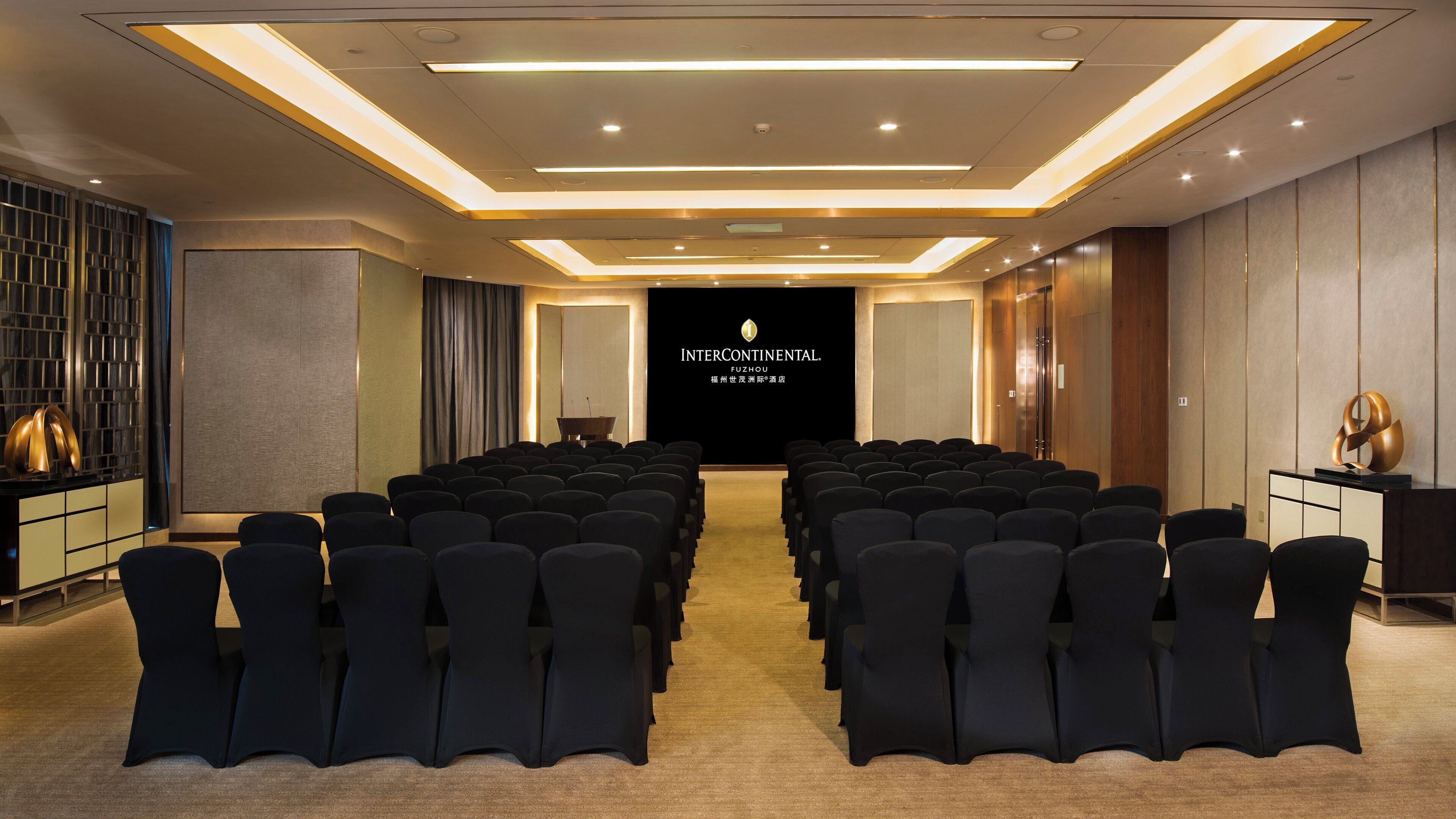 Et mødelokale med rækker af stole, der vender mod en scene, en talerstol og en stor skærm, der viser 'INTERCONTINENTAL FUZHOU'.