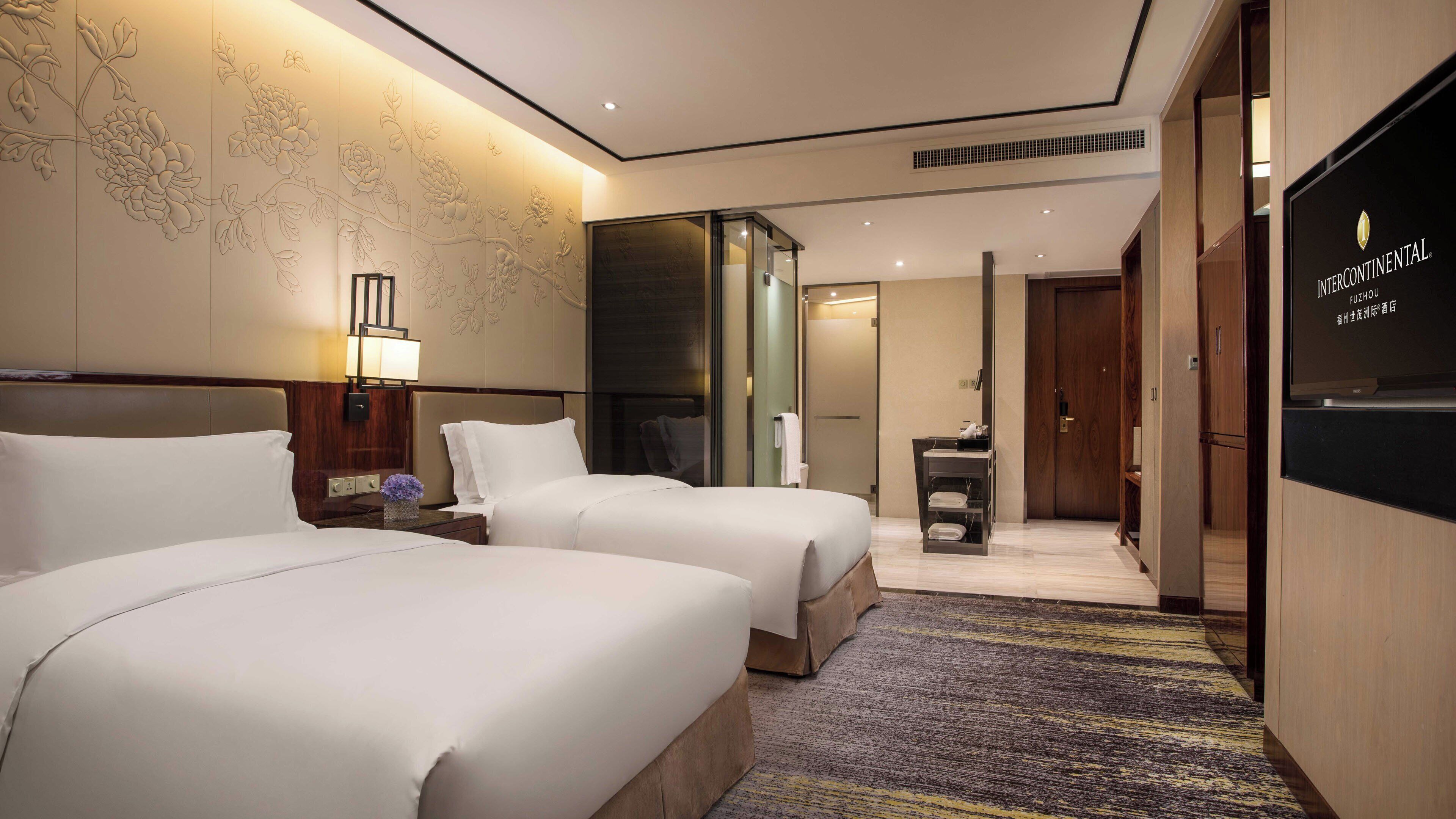 A hotel room with two beds, a nightstand, a lamp, a wall with floral patterns, a door, and a television displaying the hotel name.