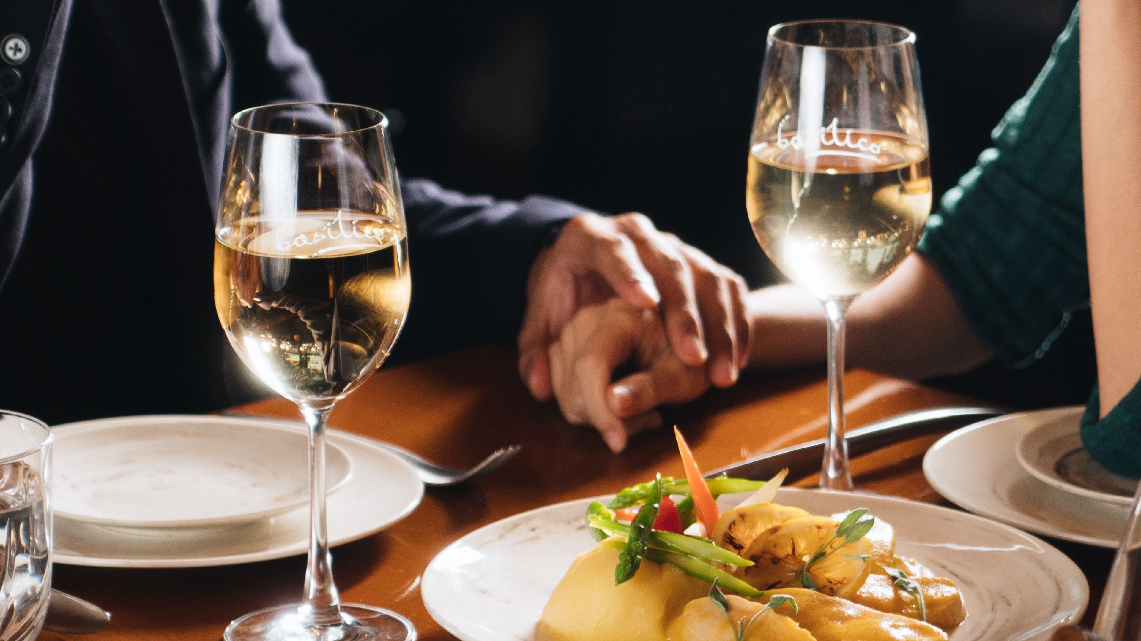 A dining table with two wine glasses, a plate of food, and a person's hands resting on the table.