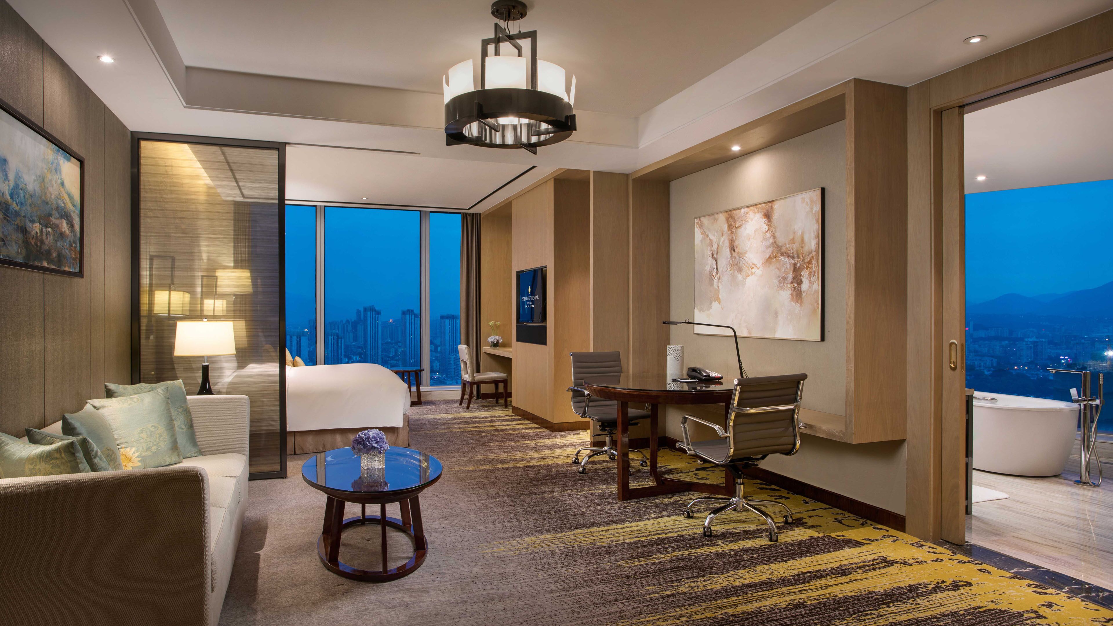 Habitación de hotel moderna con una cama grande, un escritorio, una silla y vistas a la ciudad al atardecer.