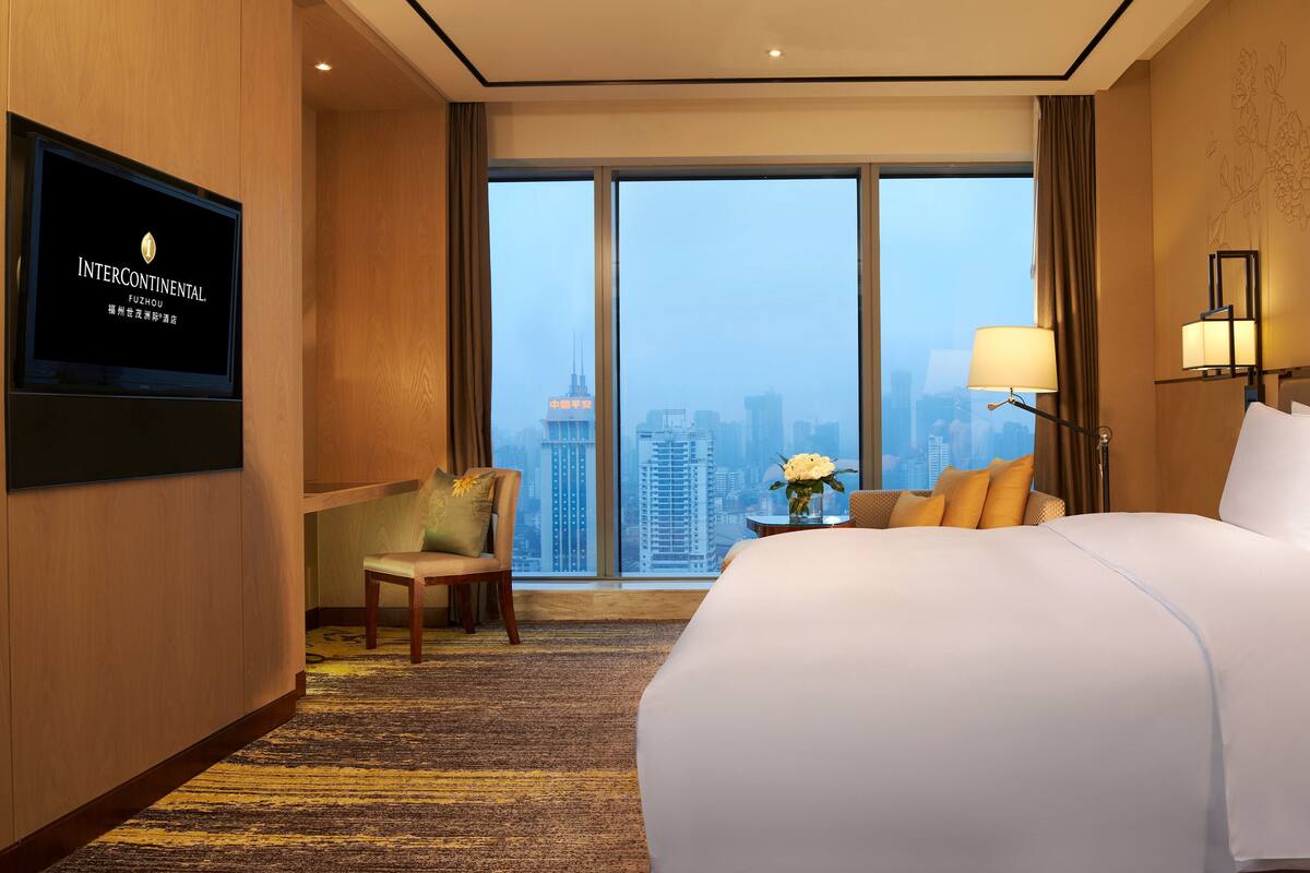 A hotel room with a large bed, a chair, a TV, and a view of the cityscape.