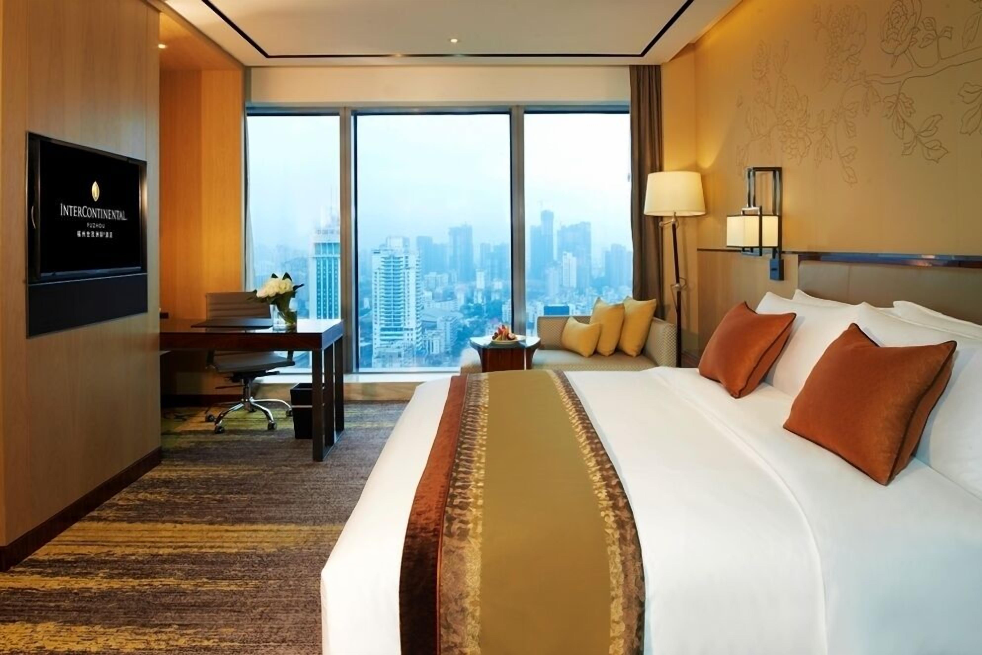 A hotel room with a large bed, a desk with a chair, a TV, and a view of the cityscape.