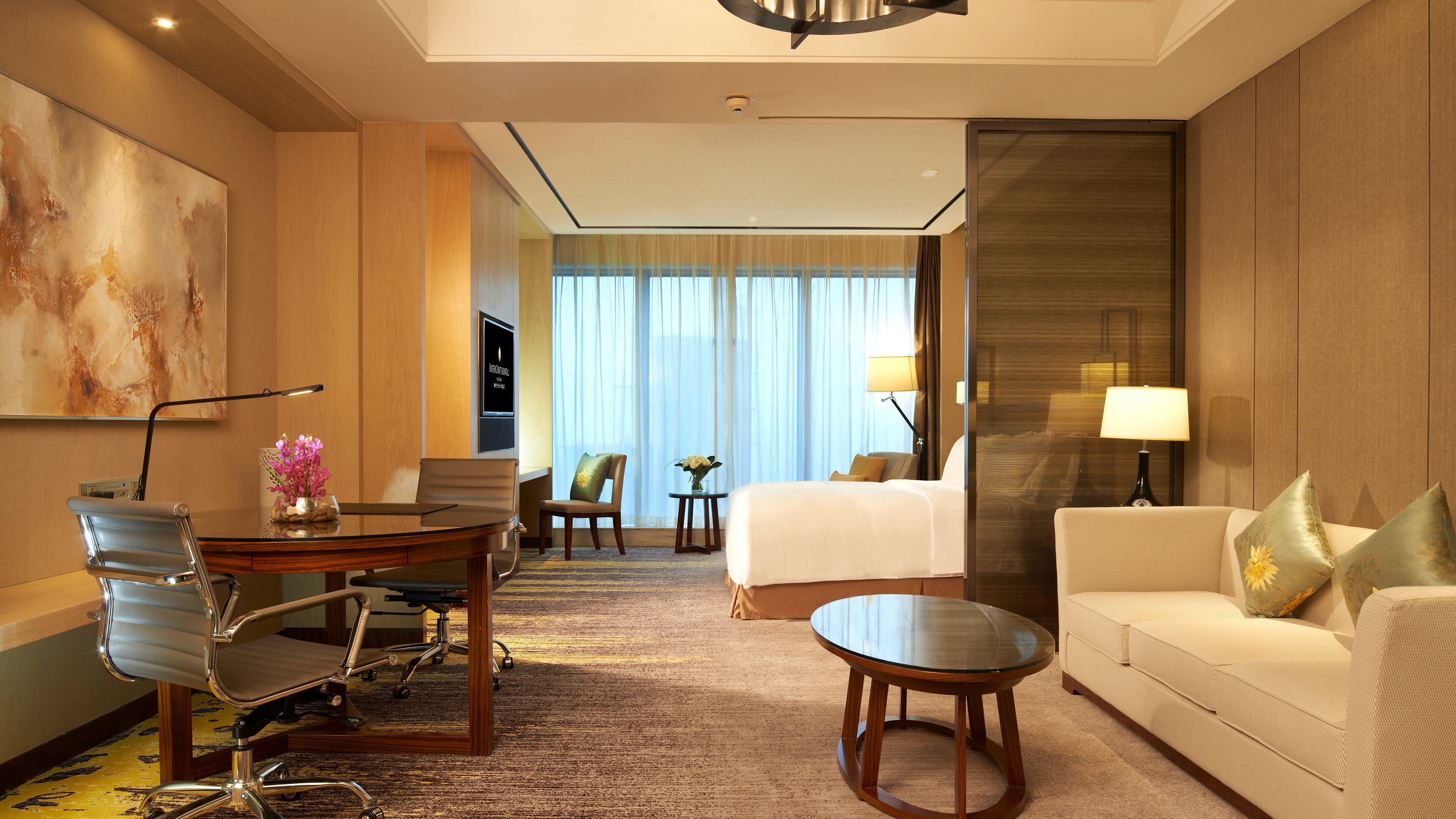A hotel room with a bed, a sofa, a desk, a chair, a lamp, and a painting on the wall.