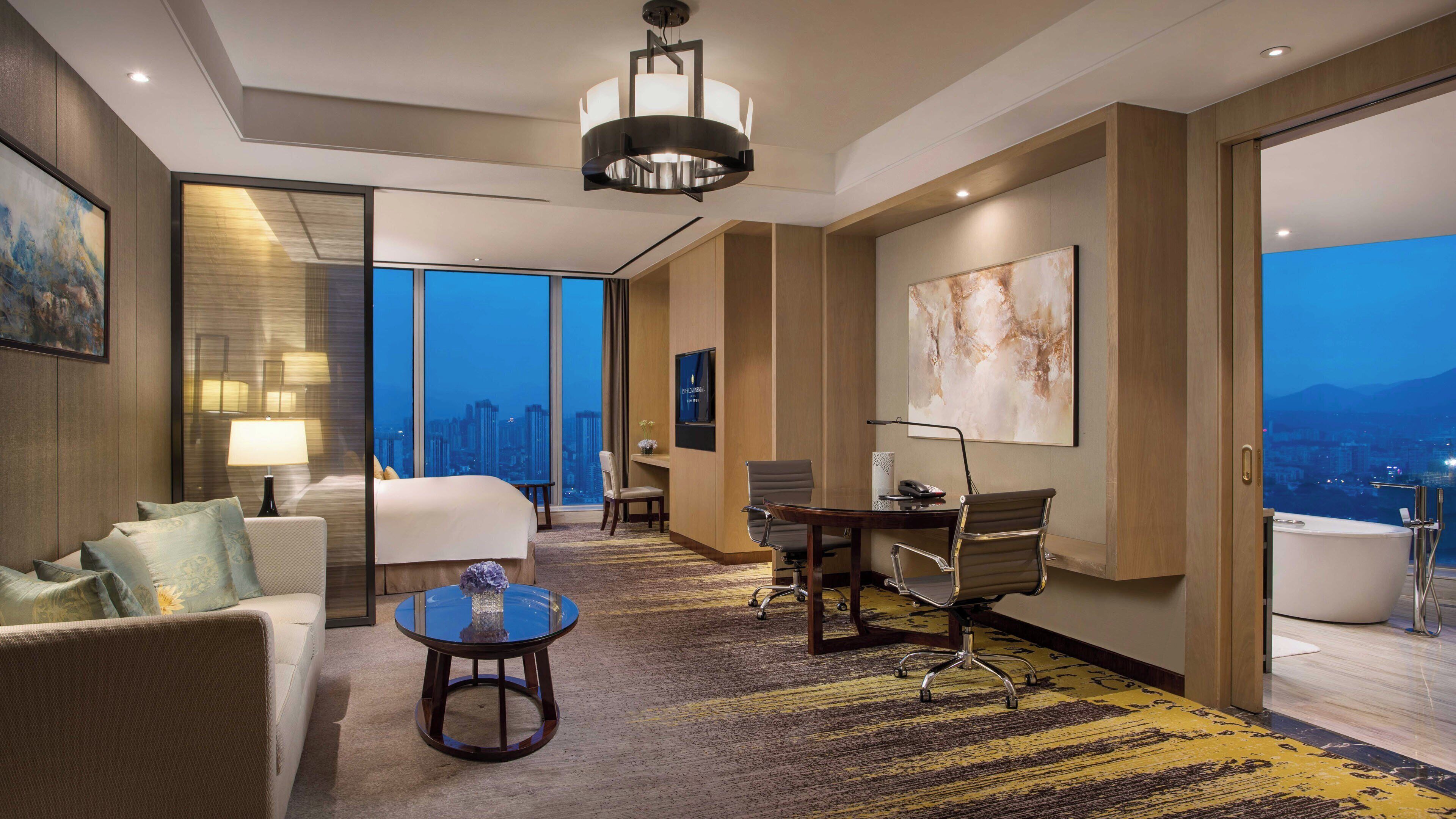 A hotel room with a bed, seating area, desk, and a large window offering a city view.