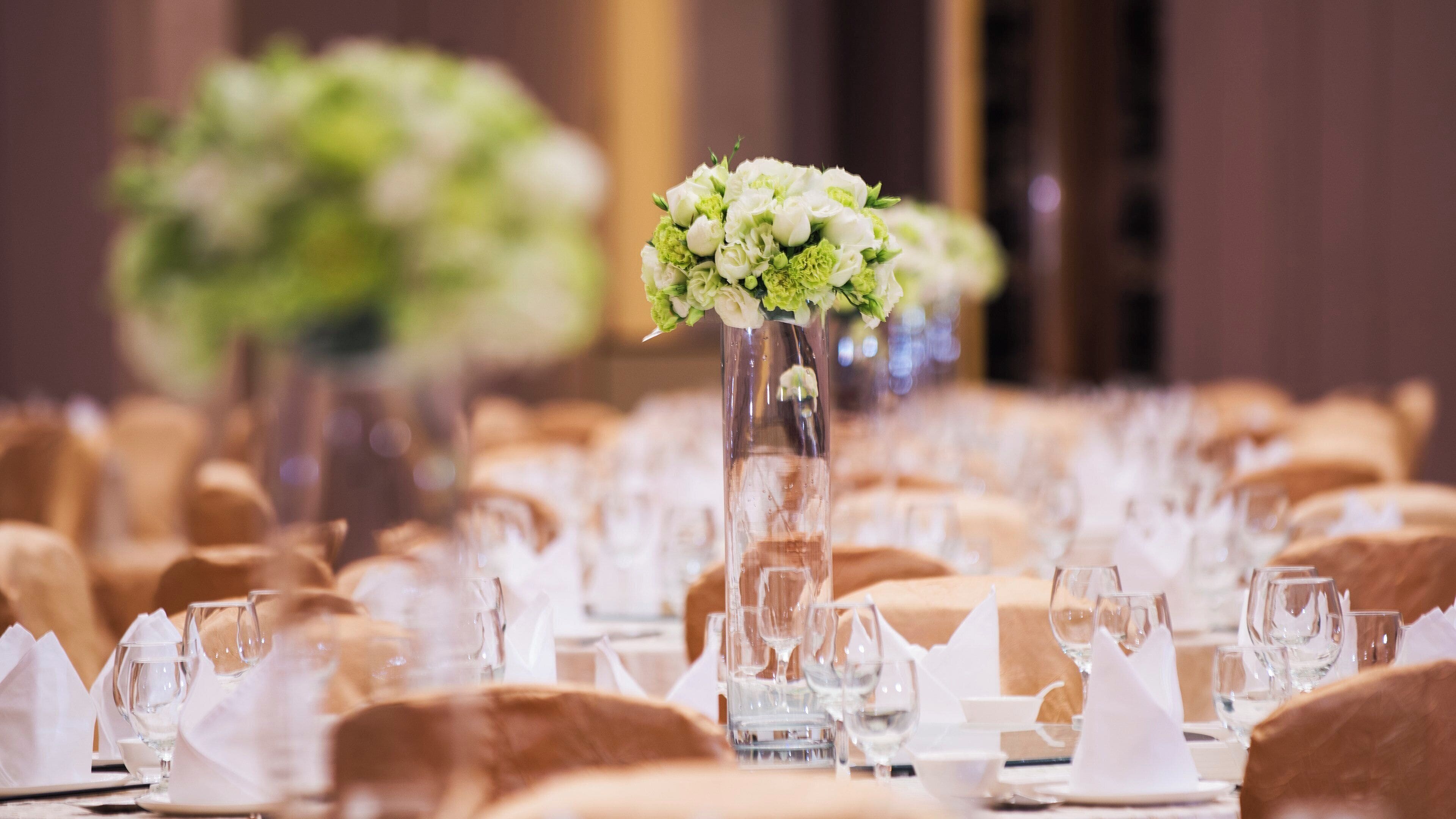 A banquet table set with white napkins, clear glassware, and a centerpiece with white flowers in a tall vase.