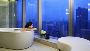 Une salle de bain moderne avec une baignoire, une petite table sur laquelle est posé un vase de fleurs, et une vue sur le paysage urbain à travers de grandes fenêtres.