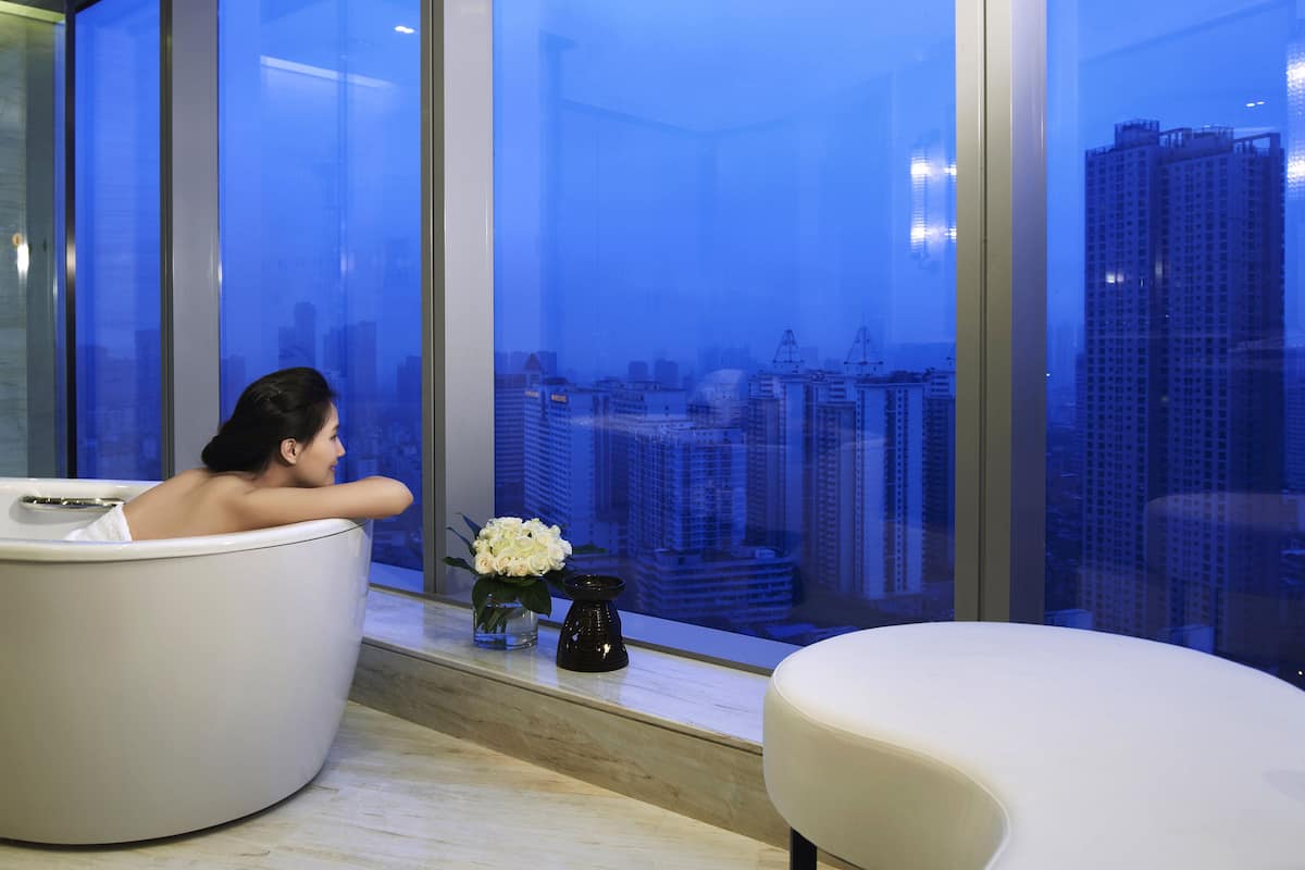 A modern bathroom with a bathtub, a small table with a vase of flowers, and a view of a cityscape through large windows.