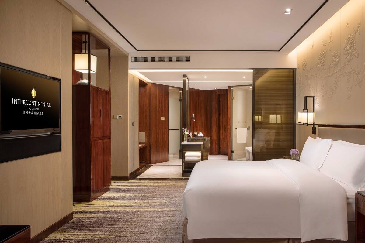 A hotel room with a bed, a TV, a desk with a lamp, and a bathroom with a mirror.