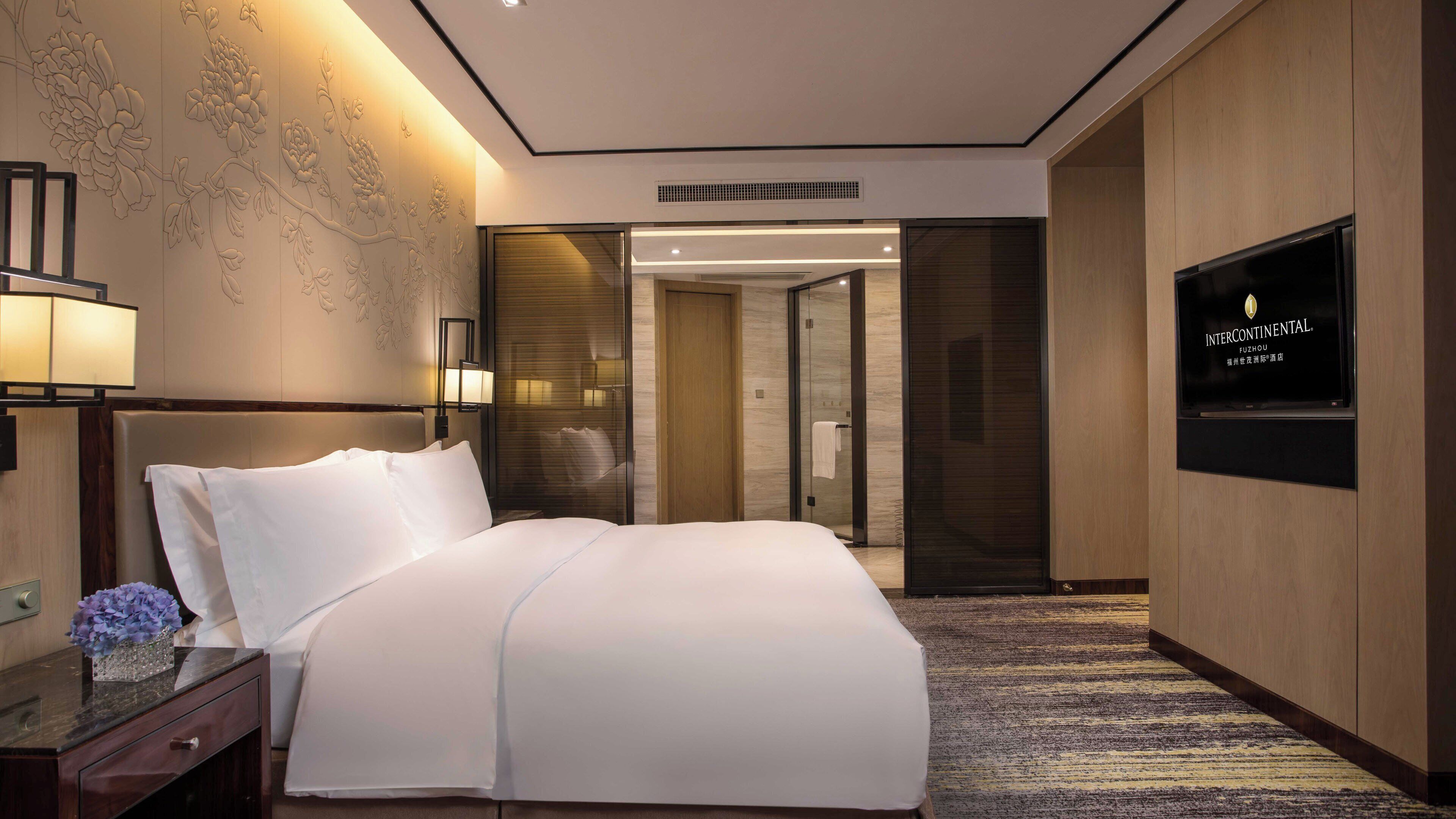 A hotel room with a large bed, a bedside table with a vase of flowers, a flat-screen TV mounted on the wall, and a carpeted floor.