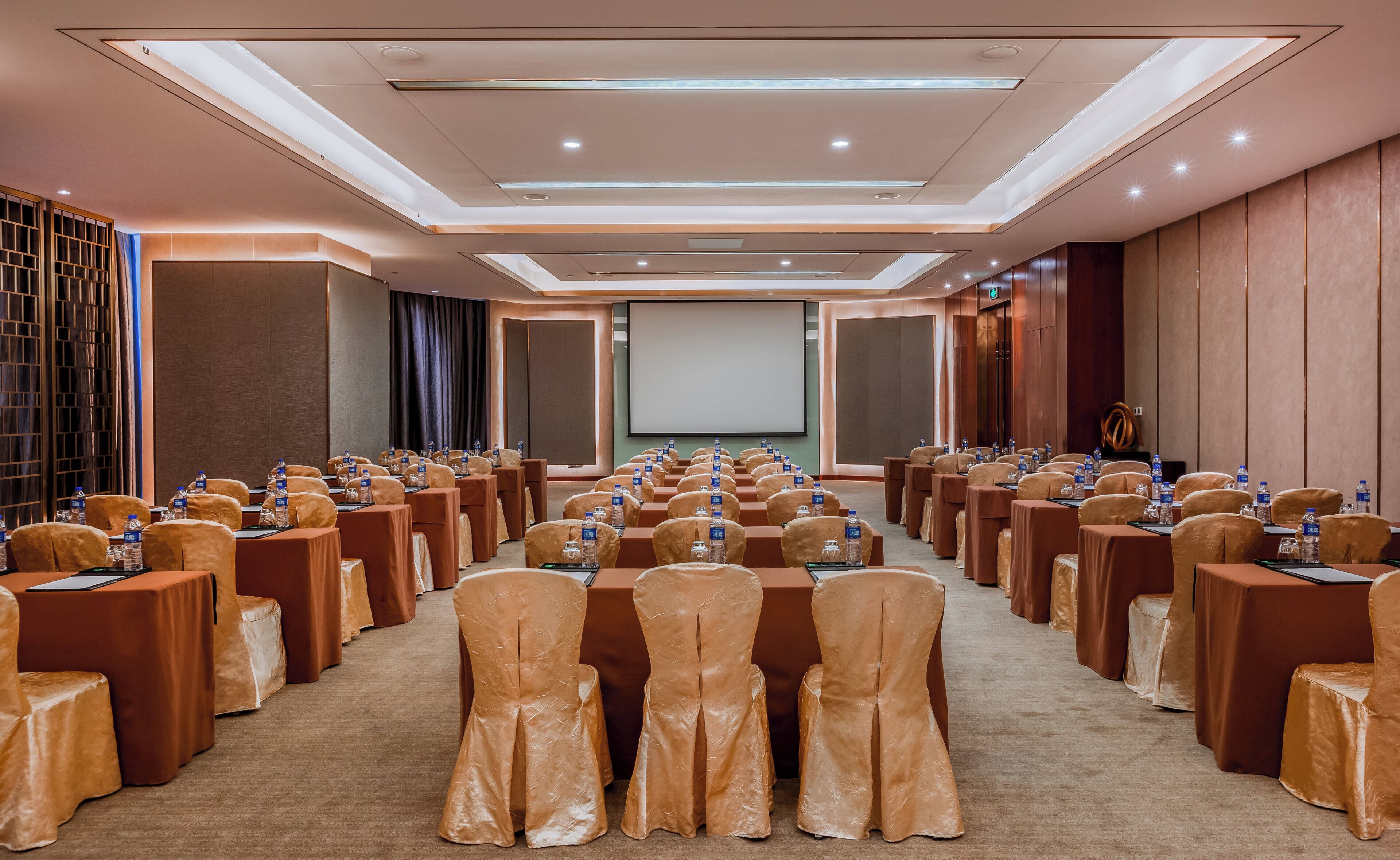 A conference room with rows of tables and chairs, a projector screen, and a carpeted floor.