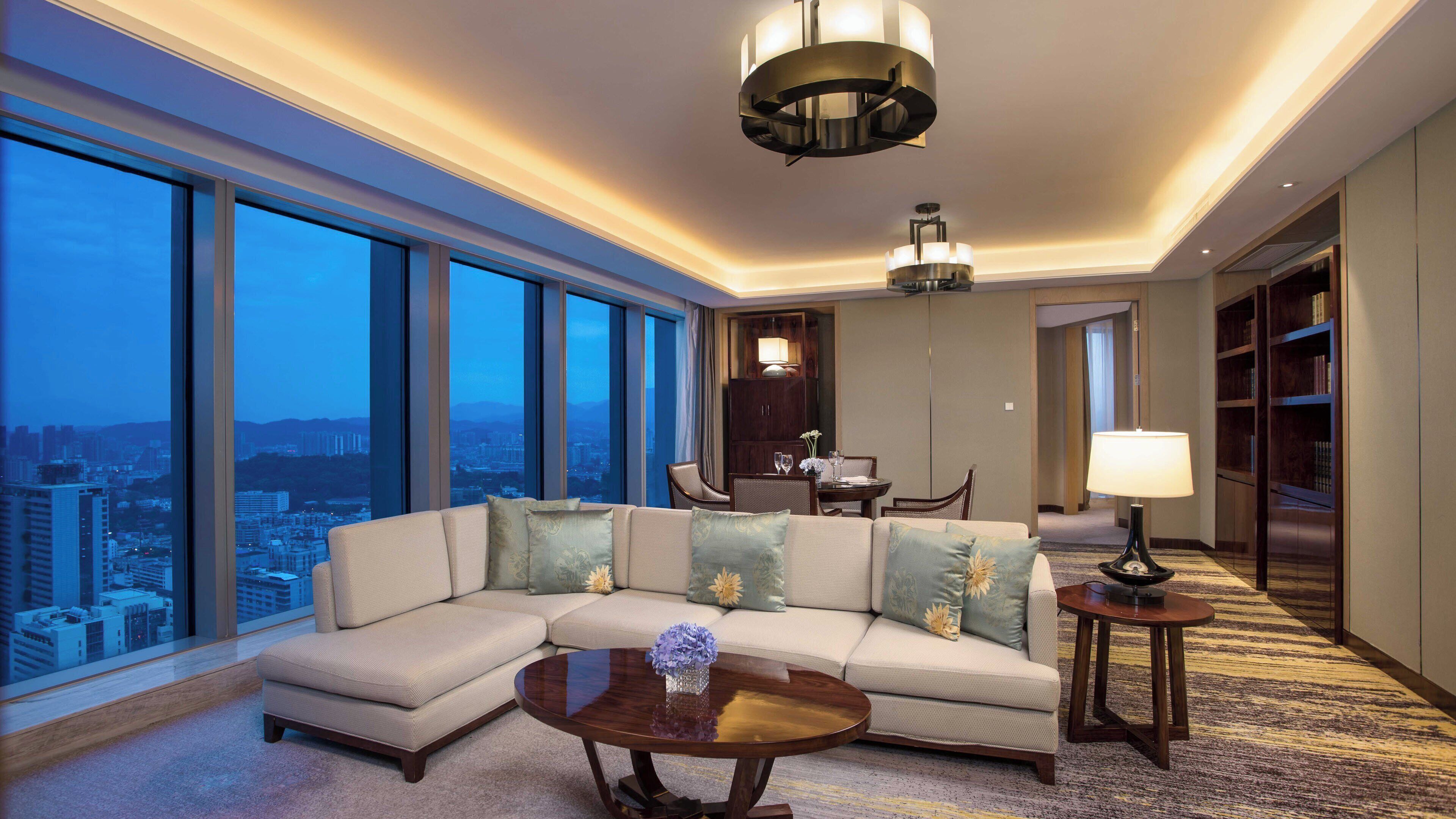 A modern hotel room with a large window, a sectional sofa, a round coffee table, and a dining area.