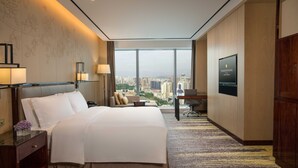 Une chambre d’hôtel avec un grand lit, un bureau avec une chaise, une télévision et une vue sur la ville grâce à de grandes fenêtres.
