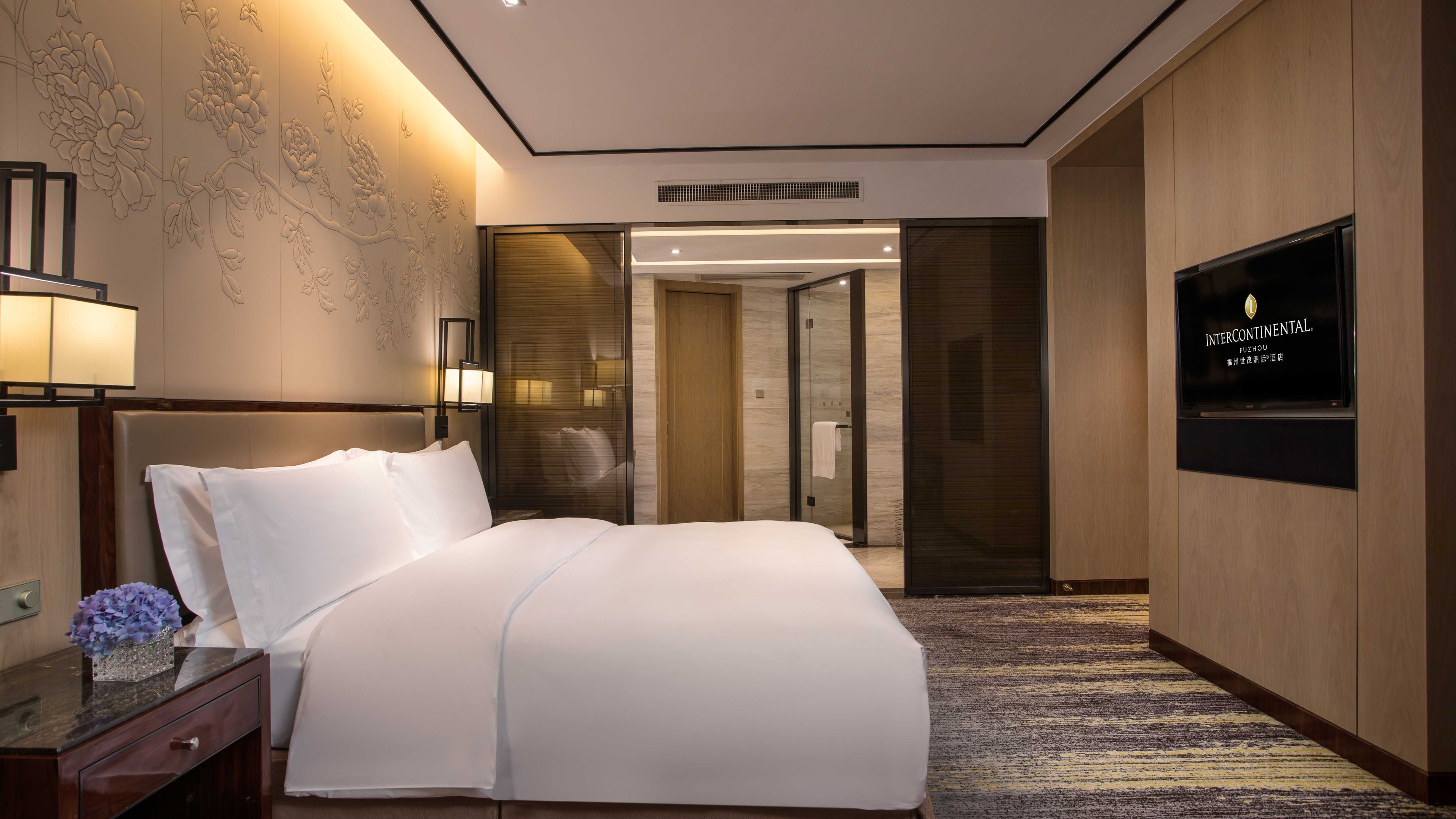 Una habitación de hotel con una cama grande, una mesita de noche con un jarrón de flores, un televisor montado en la pared y un área de ducha.