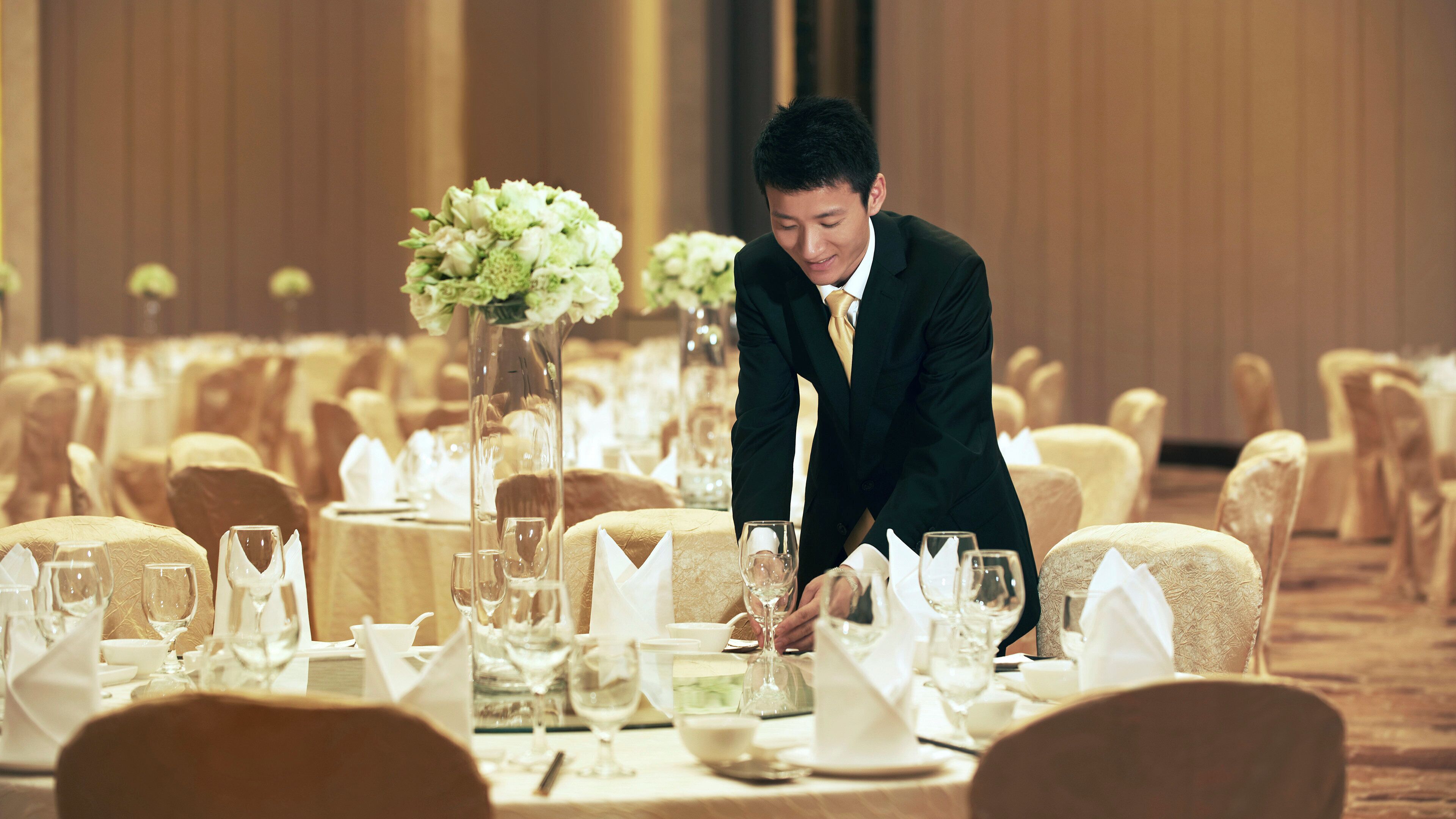 A banquet hall with round tables set for a formal event, each with white tablecloths, glassware, and floral centerpieces.