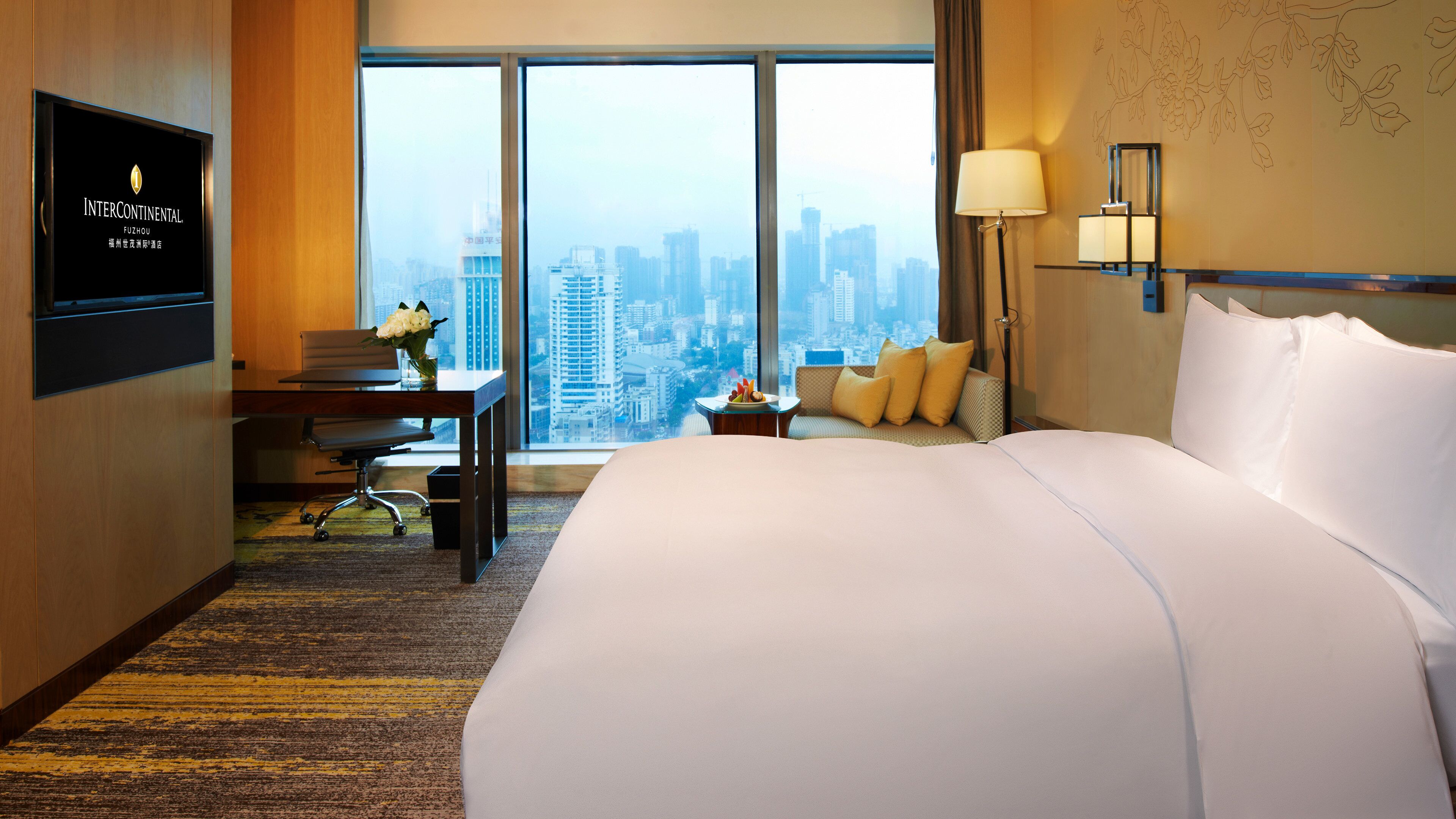 A hotel room with a large bed, a desk with a chair, a small table with fruit, a sofa with cushions, a TV on the wall, and a city view through large windows.
