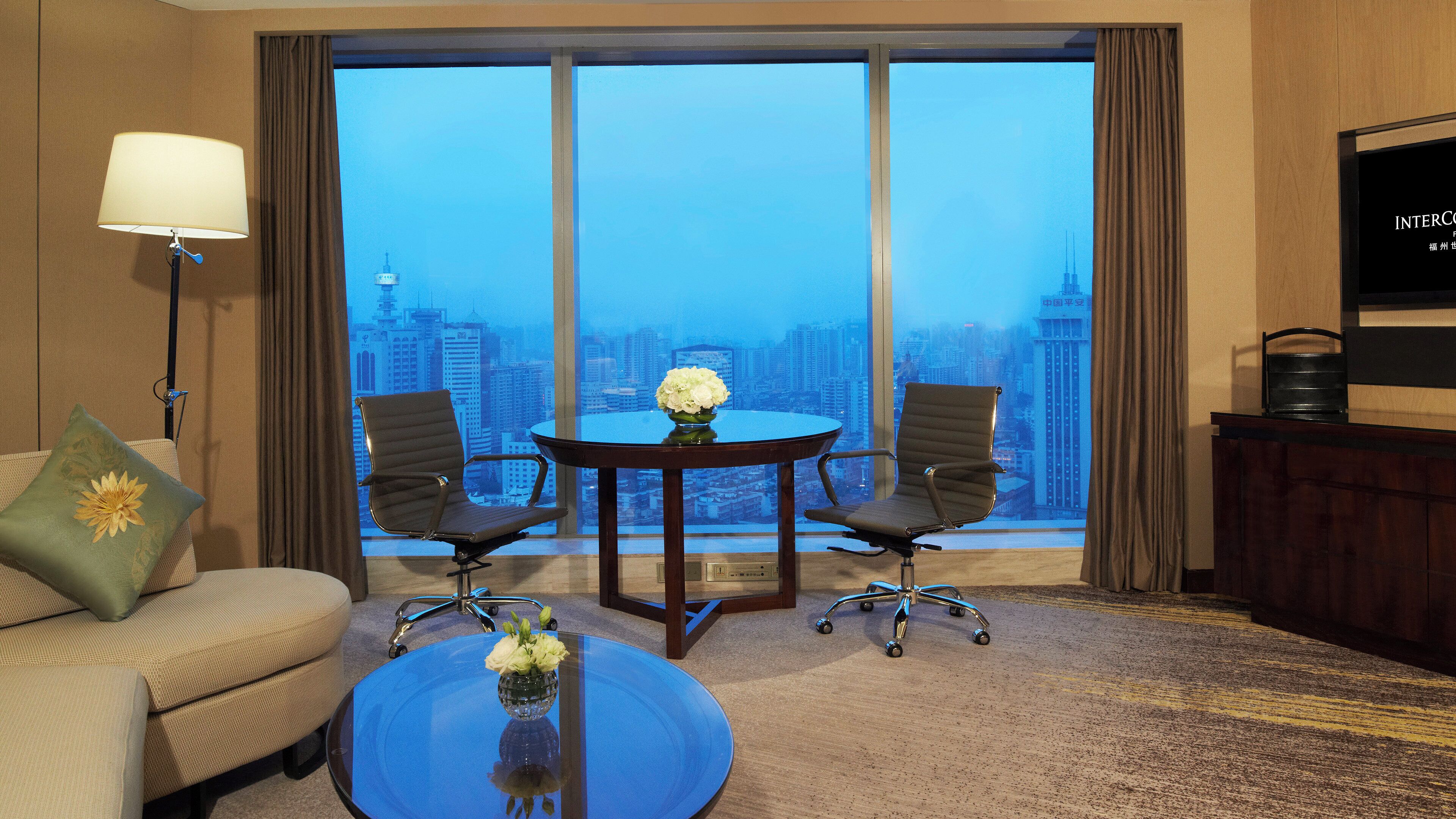 A hotel room with a city view, a round table with two chairs, a sofa, a coffee table with a vase, and a lamp.