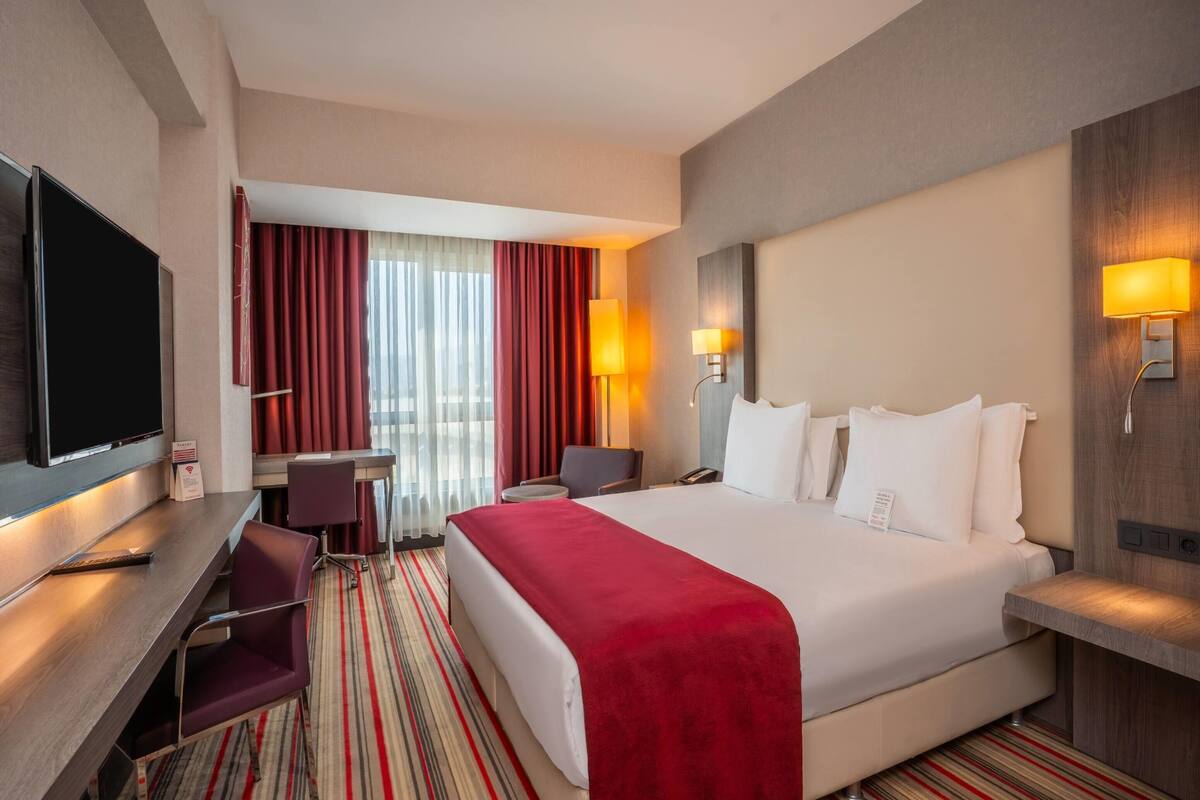 Een hotelkamer met een groot bed, een bureau met een televisie, een stoel, een bureaulamp en een raam met gordijnen.