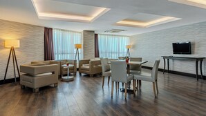 Habitación de hotel moderna con sofá, mesa de comedor y sillas, televisión de pantalla plana y un ventanal con cortinas.