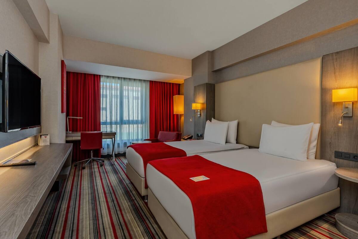 Een hotelkamer met twee bedden, een bureau, een televisie en een raam met rode gordijnen.