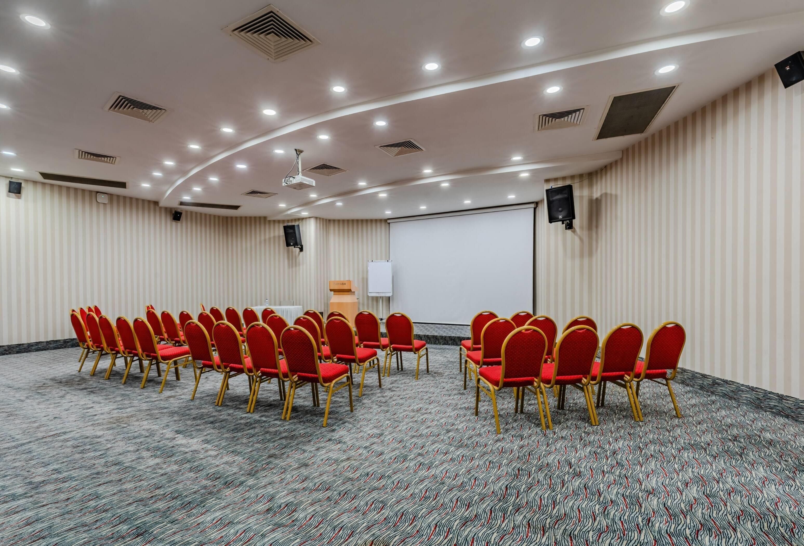 A conference room with rows of red chairs, a whiteboard, and a projector screen.